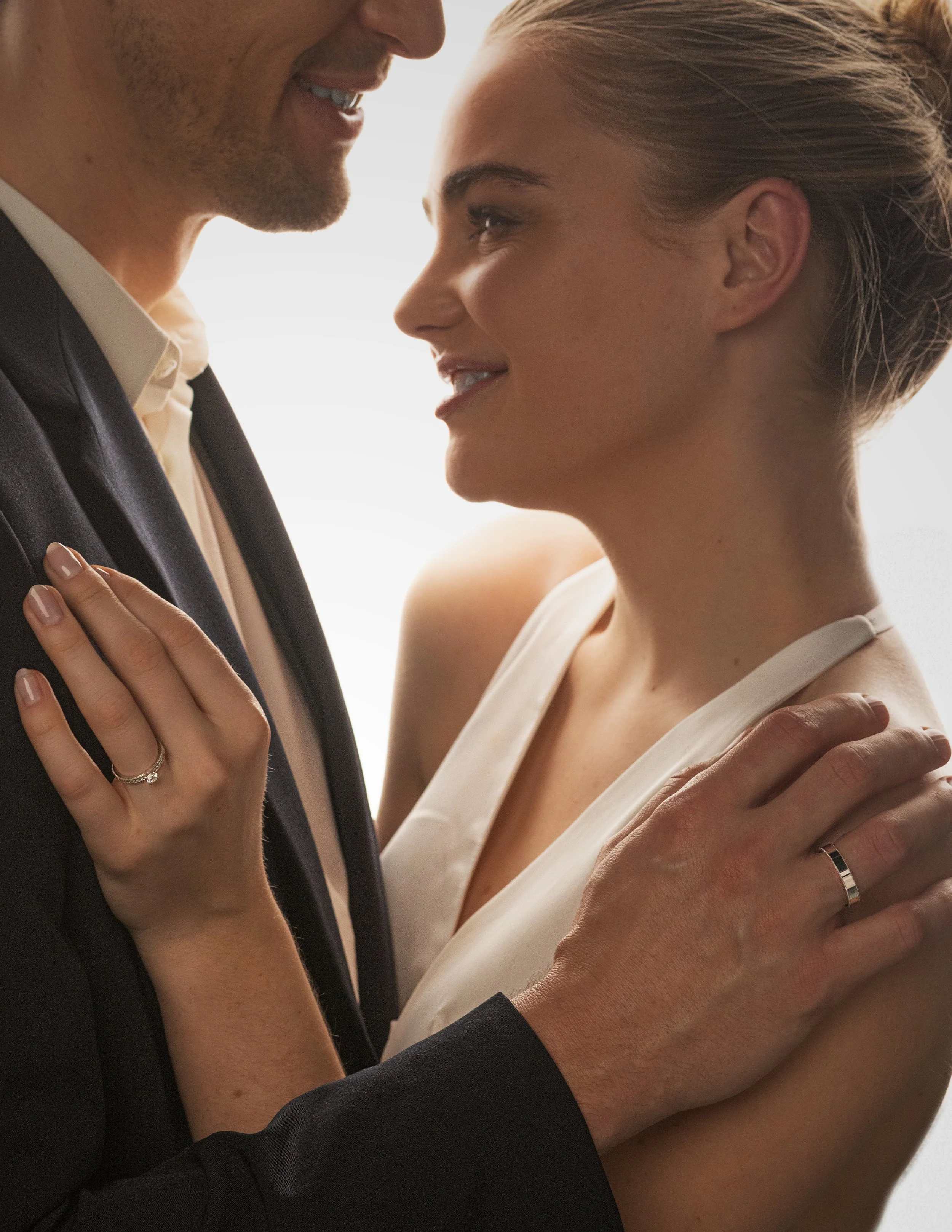 Ein Paar lächelt sich an; beide tragen Eheringe. Die Frau trägt ein weißes Kleid, und der Mann einen dunklen Anzug.