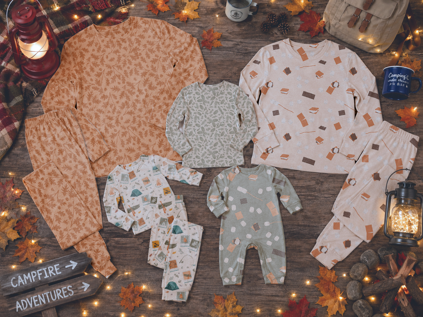 Flat lay of matching pajamas for adults and children with autumn-themed patterns, surrounded by fall leaves, lanterns, a camping mug, and a snack bag, on a wooden surface.