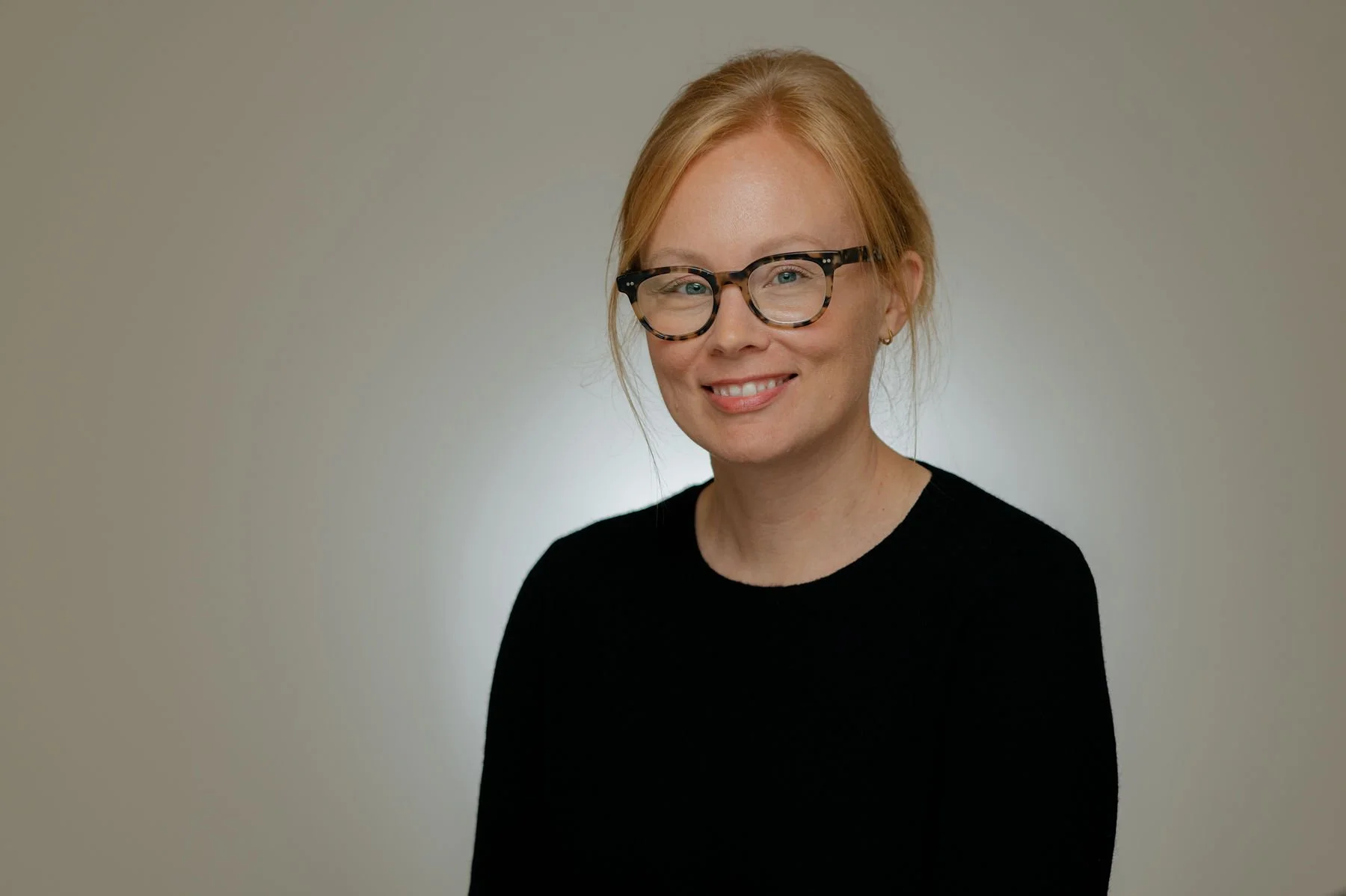 A woman with red hair, glasses, and a black shirt smiling against a plain light-colored background.