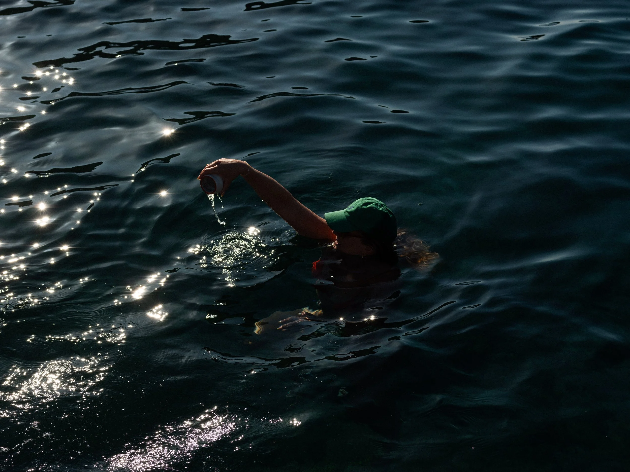 A person in the water, wearing a green hat, is reaching out and holding a small container. The water is dark and reflective with sunlight creating bright spots on the surface.