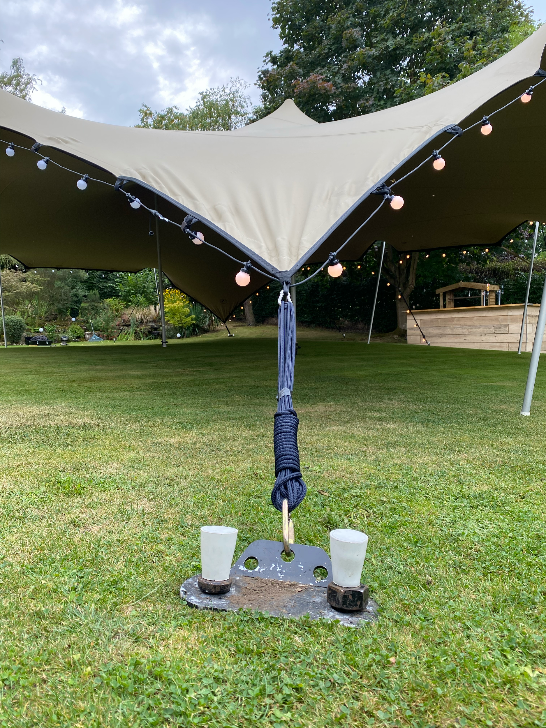 A beige canopy tent with string lights, set up in a grassy yard with trees and a wooden fence. In the foreground, there is a metal umbrella base with two white plastic cups on black bases, and a rolled-up umbrella or outdoor heater.