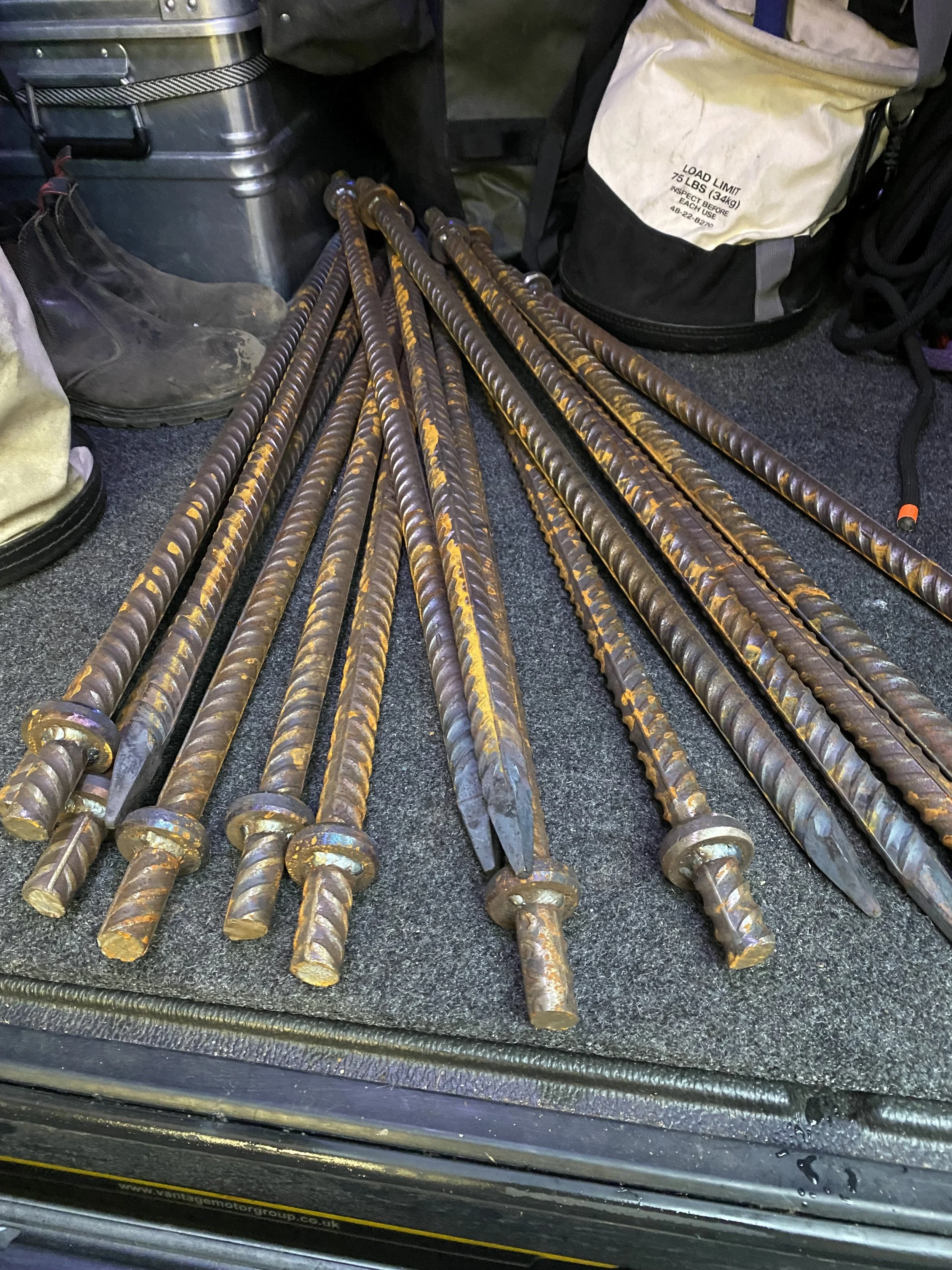 Multiple steel rebar rods with spiral ridges and flat tips laid on a black surface, surrounded by a backpack, boots, and a metallic container.