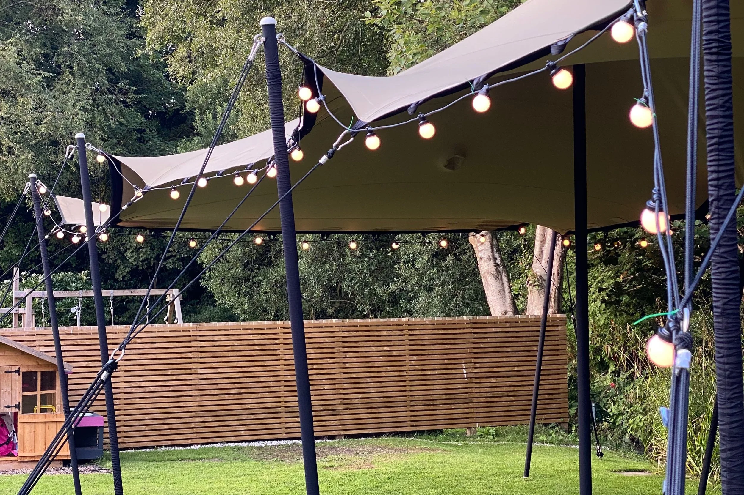 Outdoor backyard with a large beige canopy, string lights hanging, green grass, wooden fence, trees, a small wooden shed, and part of a trampoline in the foreground.