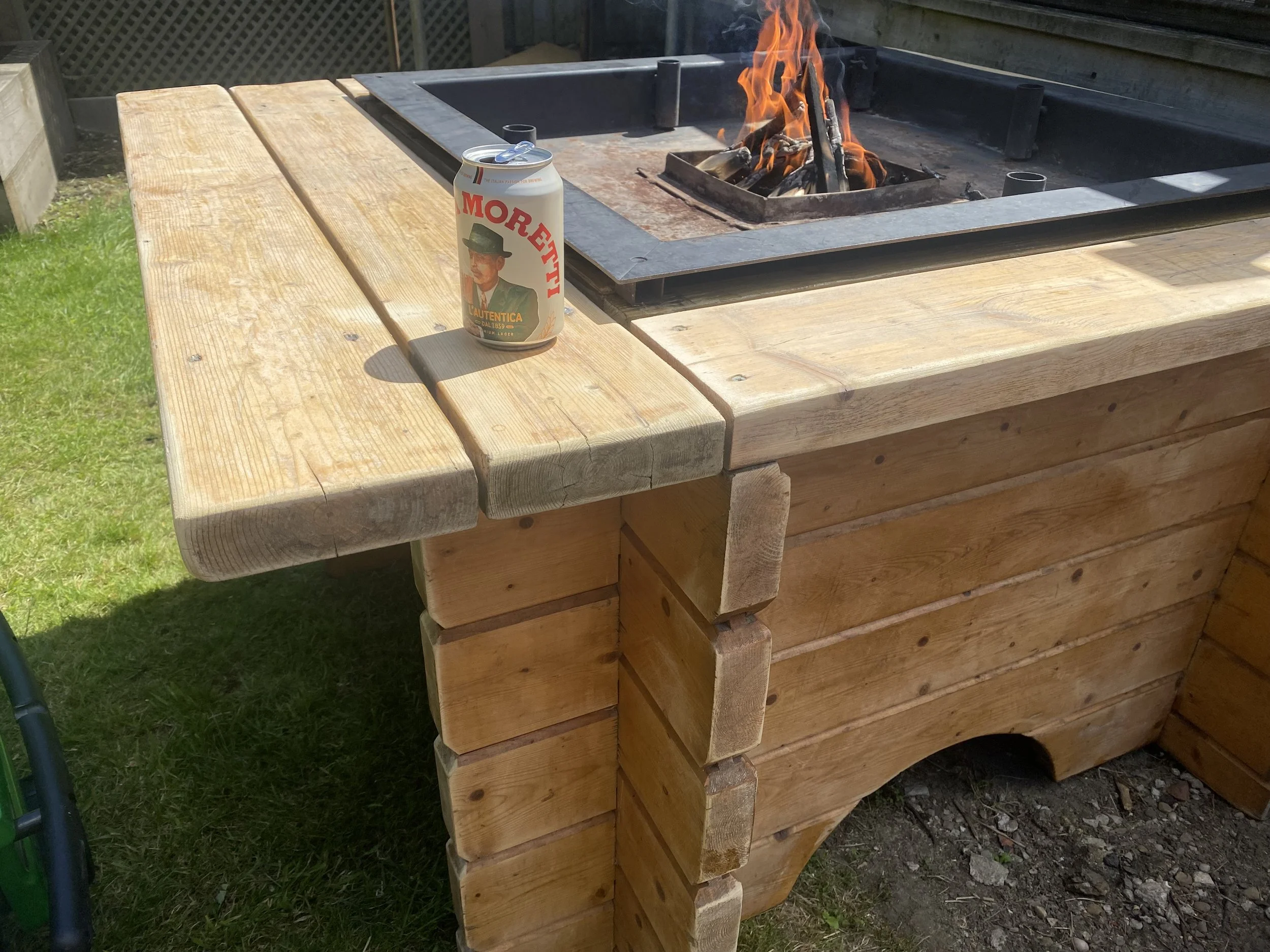 A wooden outdoor grill with an open fire, a can of beer with a vintage-style label, and a grassy yard in the background.