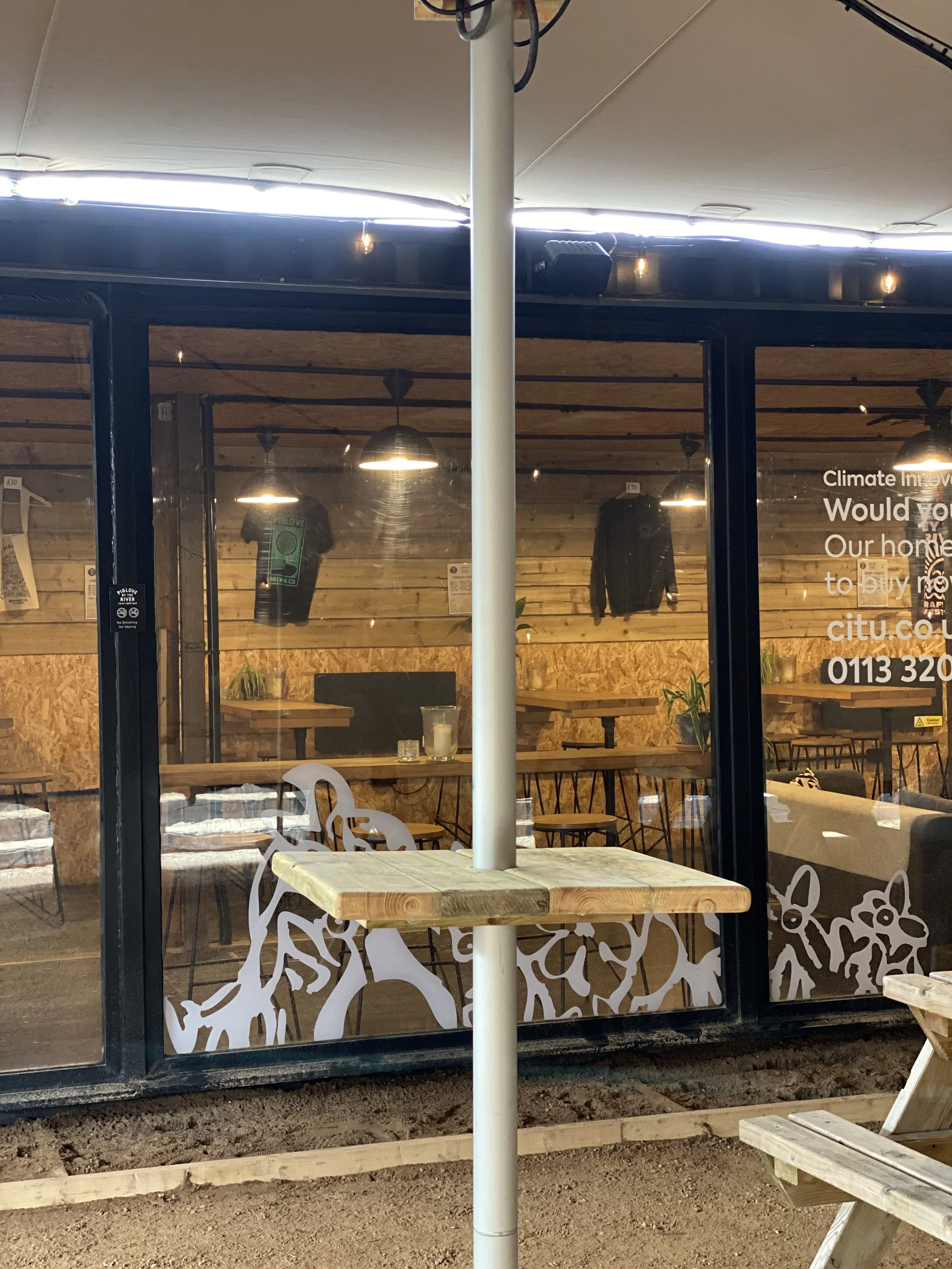 View of the outside of a modern cafe with glass walls, wooden interior, black chairs, and hanging lights. A wooden mountains with white dog illustrations is on the glass. Outer table with a parasol pole, dirt ground, and wooden bench.