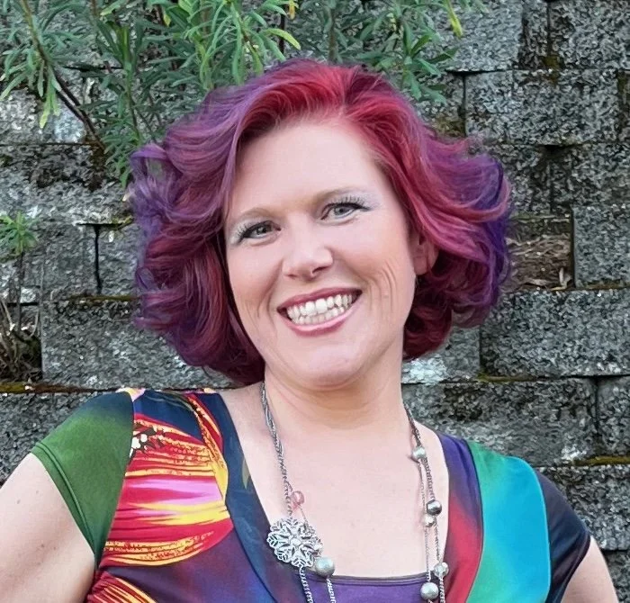 A smiling woman with curly hair dyed in shades of pink and purple, wearing a colorful dress and a necklace, standing outdoors in front of a stone wall with green foliage.