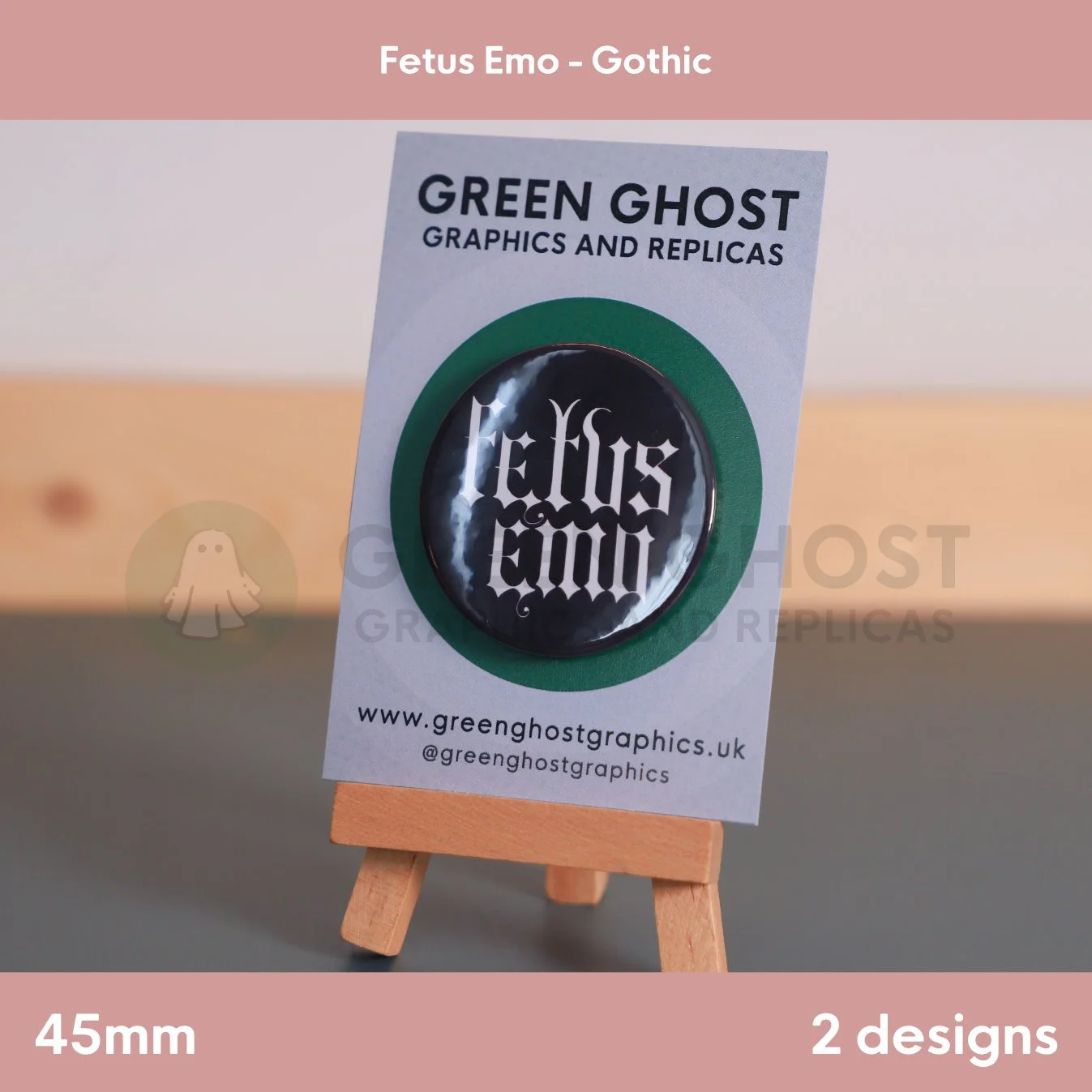 A round black badge with white Gothic text "Fetus Emo" displayed on a branded backing card on a wooden easel.
