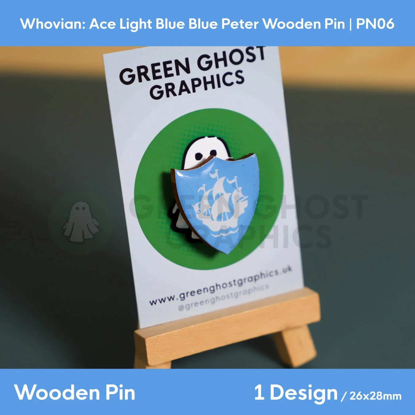 A blue shield‑shaped wooden pin displaying a white sailing ship motif mounted on a Green Ghost Graphics card on a small wooden easel.