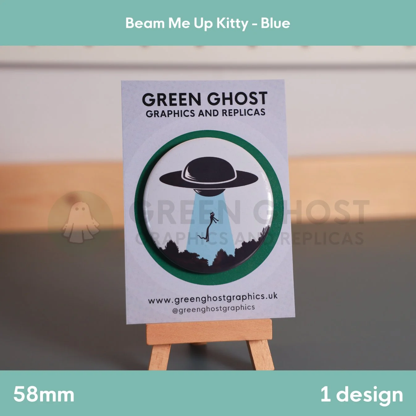 A round badge with a blue UFO beam lifting a cat displayed on a branded backing card on a wooden easel.