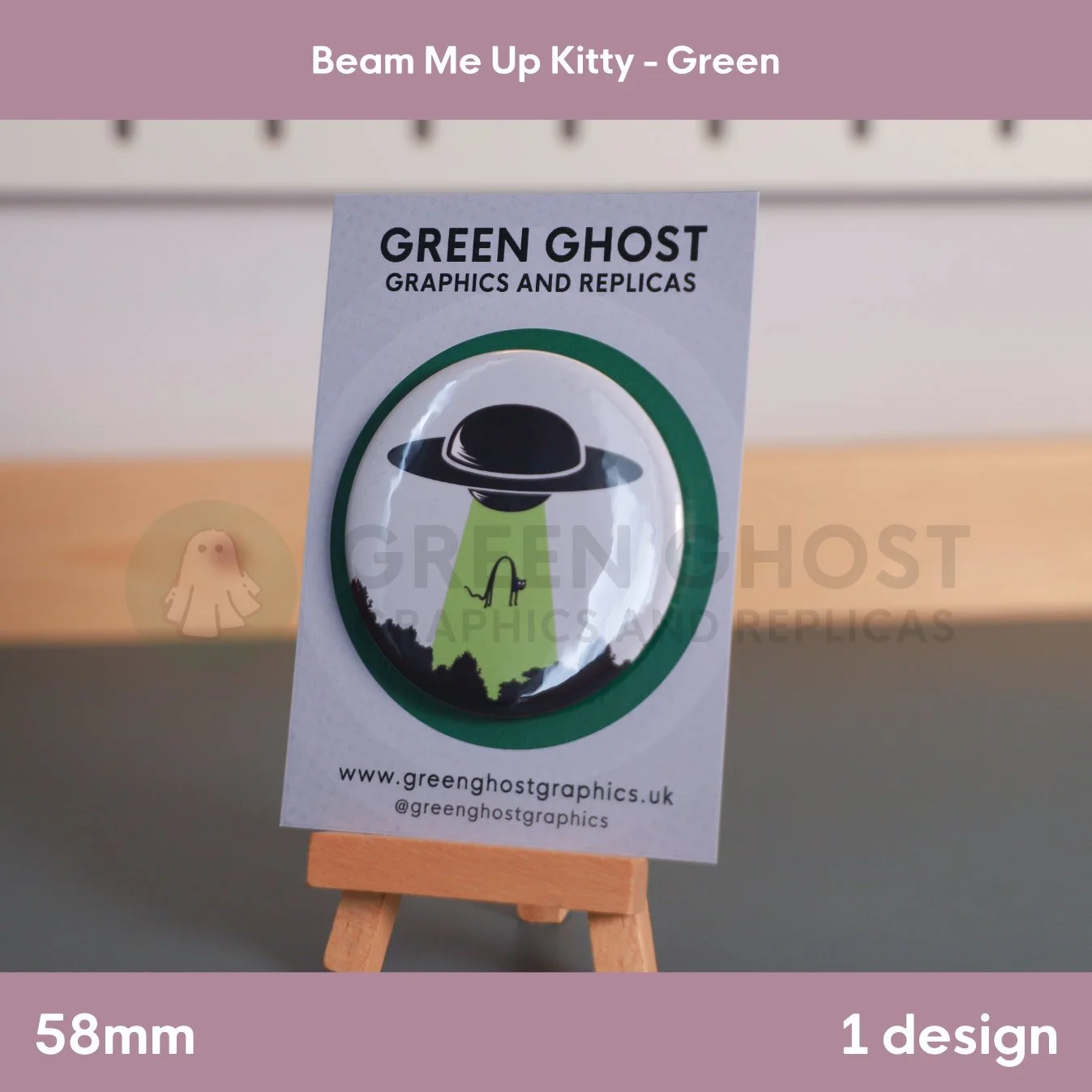 A round badge with a green UFO beam lifting a cat displayed on a branded backing card on a wooden easel.