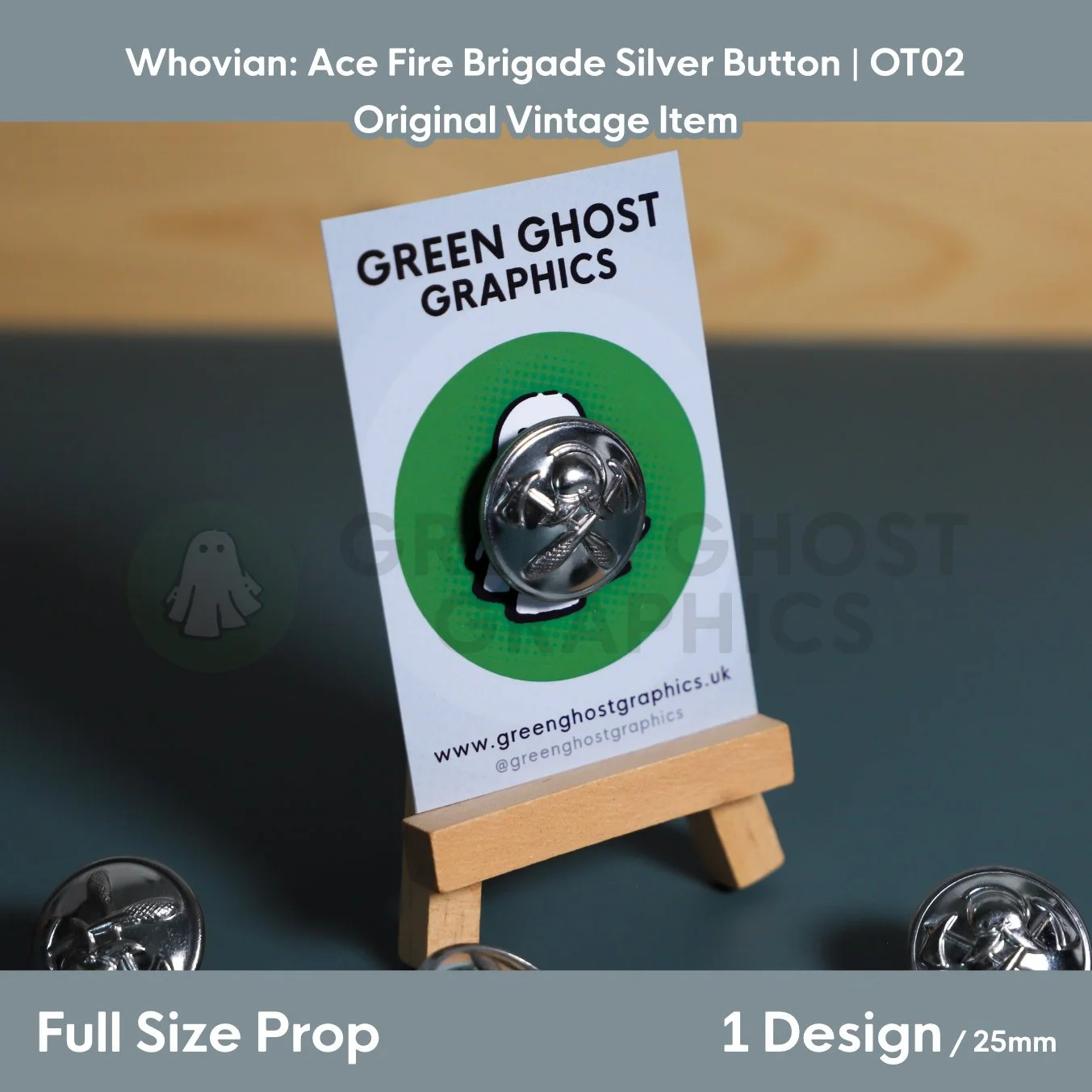A circular silver button featuring a raised fire service emblem mounted on a Green Ghost Graphics display card on a wooden easel.