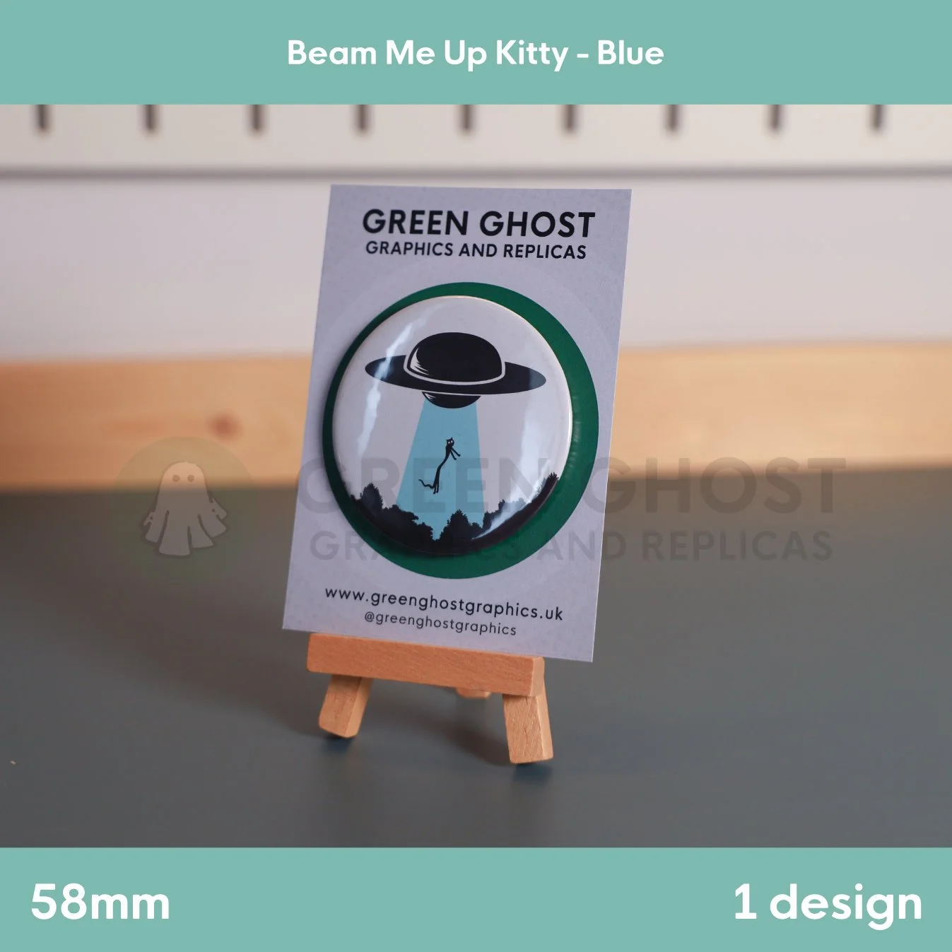 A round badge with a blue UFO beam lifting a cat displayed on a branded backing card on a wooden easel.