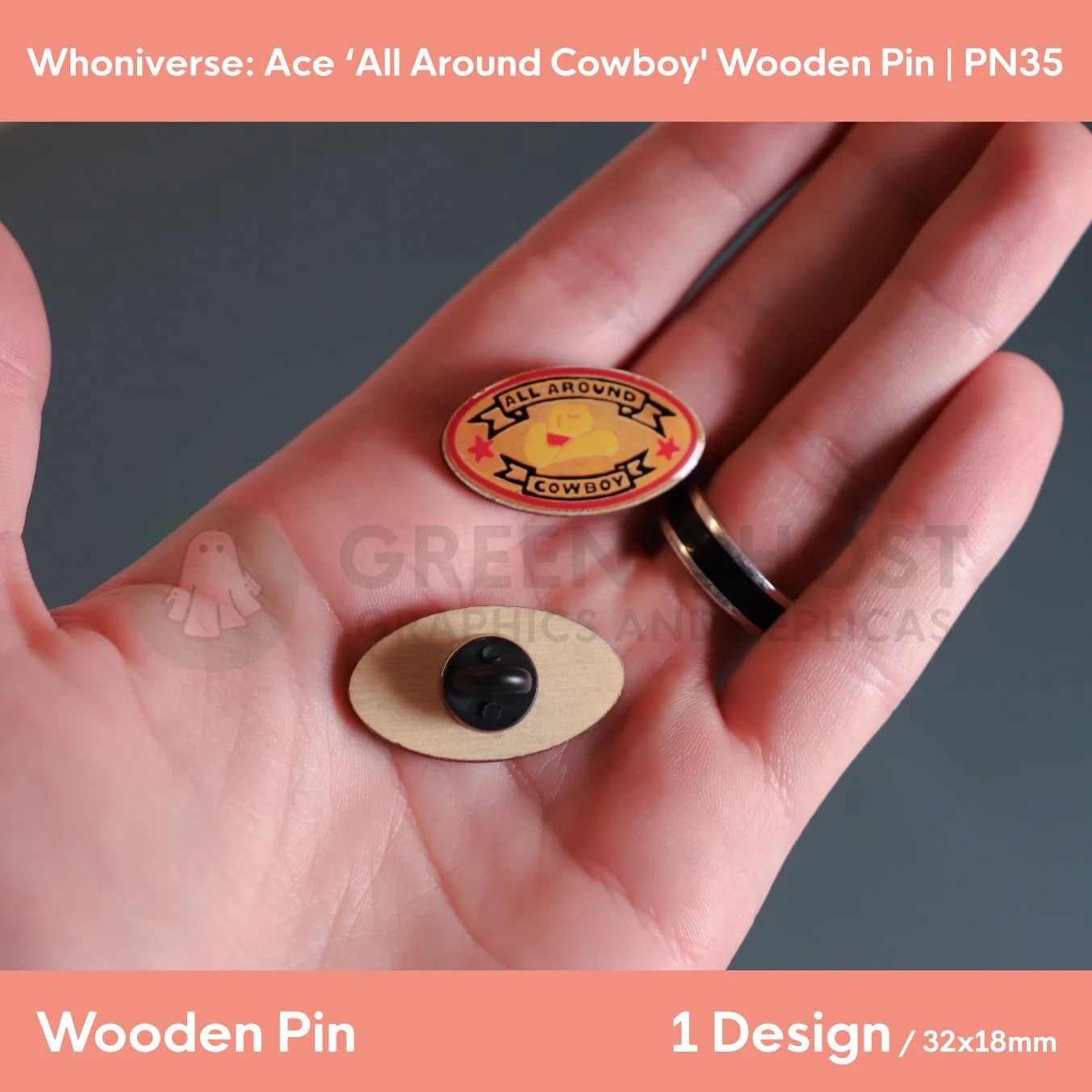 A close‑up of two oval “ALL AROUND COWBOY” wooden pins held in a hand, showing the front text design and the back with a clasp attachment.