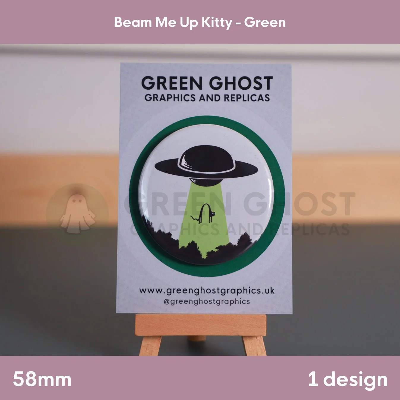 A round badge with a black ufo and green UFO beam lifting a cat displayed on a branded backing card on a wooden easel.