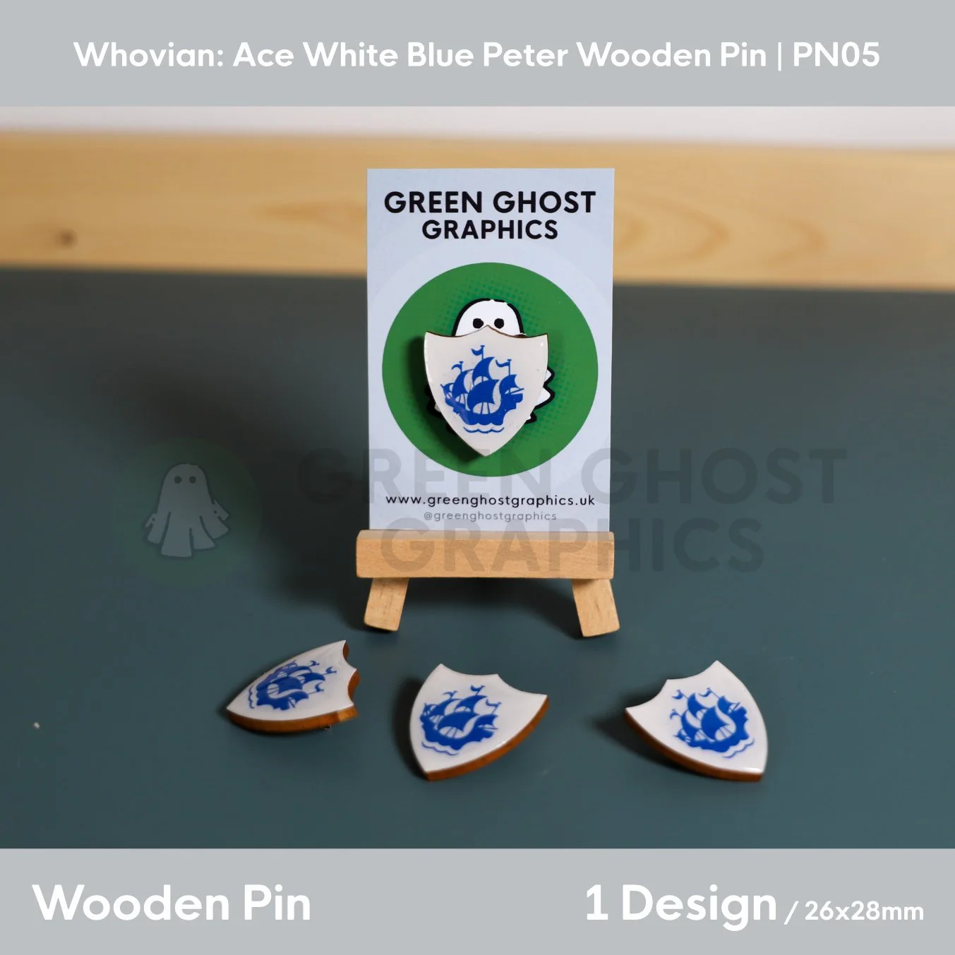A white shield‑shaped wooden pin featuring a blue sailing ship design displayed on a Green Ghost Graphics card with three matching pins arranged below.