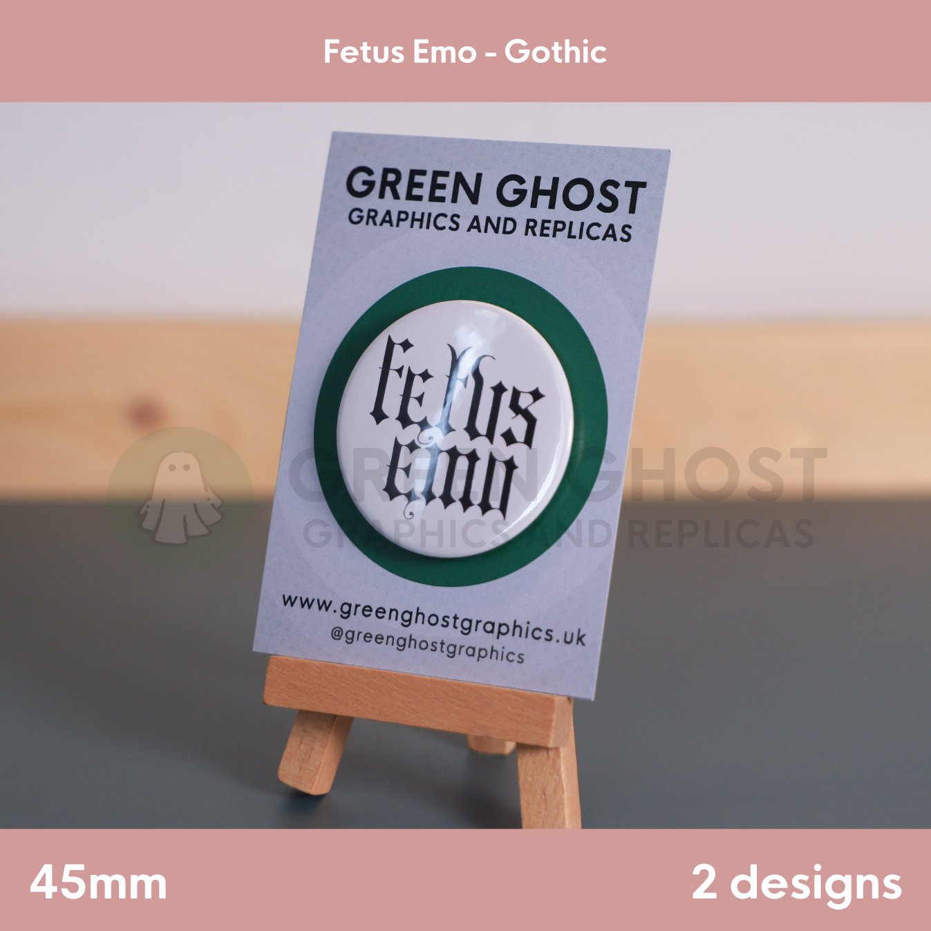 A round white badge with black Gothic text "Fetus Emo" displayed on a branded backing card on a wooden easel.