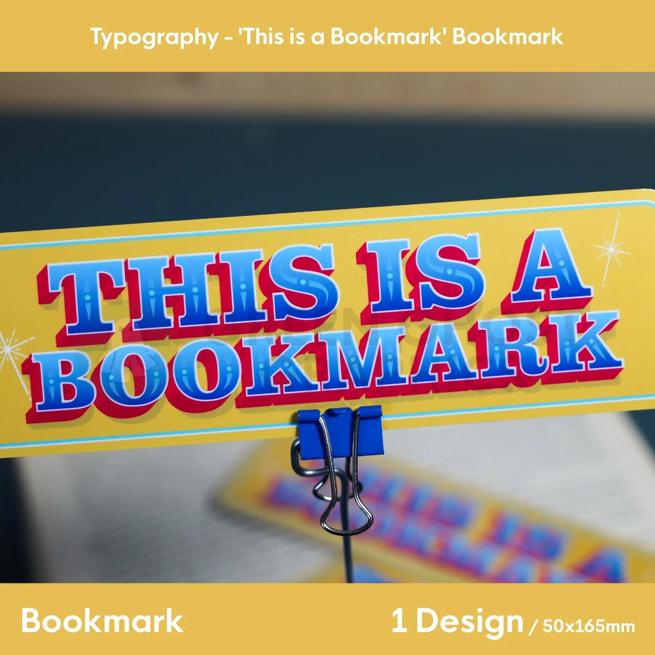 A tall yellow bookmark with colourful “THIS IS A BOOKMARK” lettering clipped upright in a blue binder clip beside an open book.