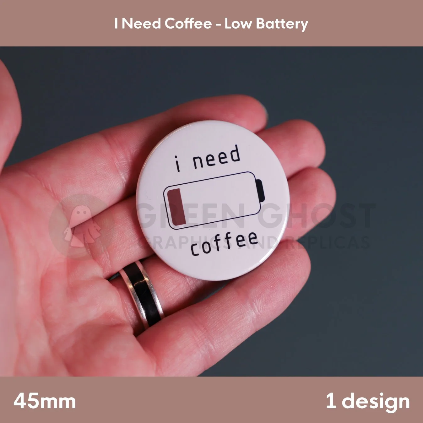 A round white badge with a low‑battery icon and the text “i need coffee” held in a hand against a grey background.
