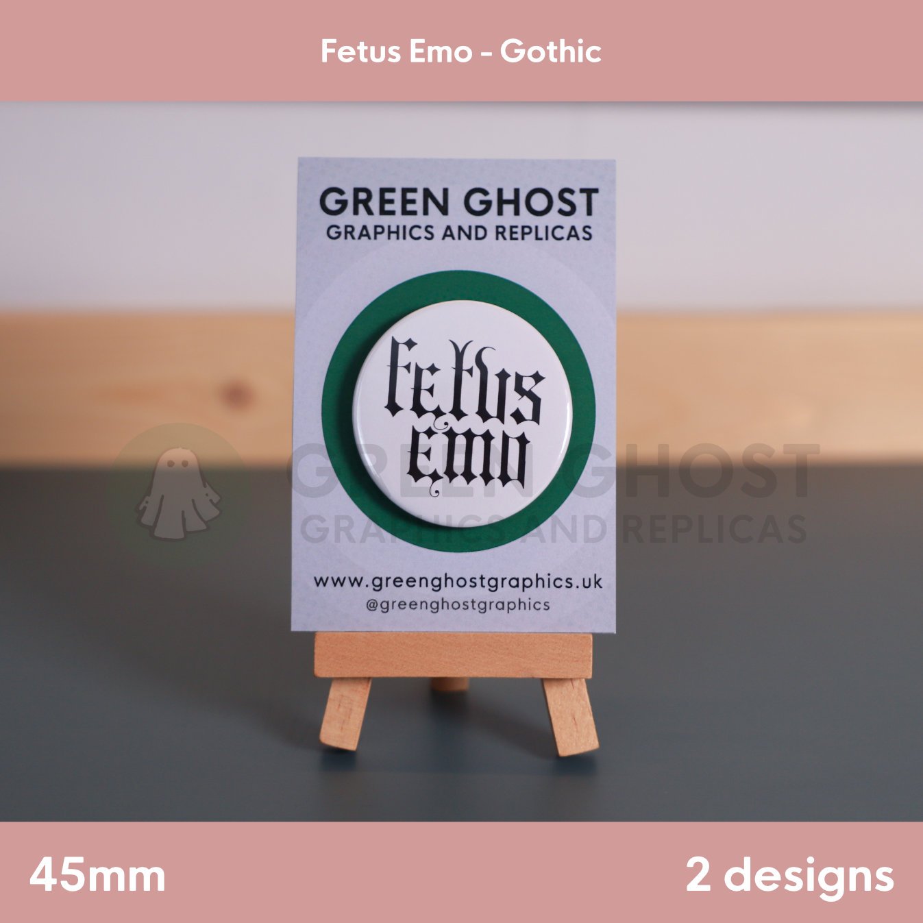 A round white badge with black Gothic text "Fetus Emo" displayed on a branded backing card on a wooden easel.