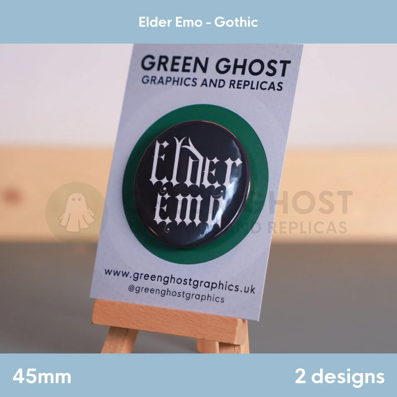 A round black badge with white Gothic "Elder Emo" text displayed on a branded backing card on a wooden easel.