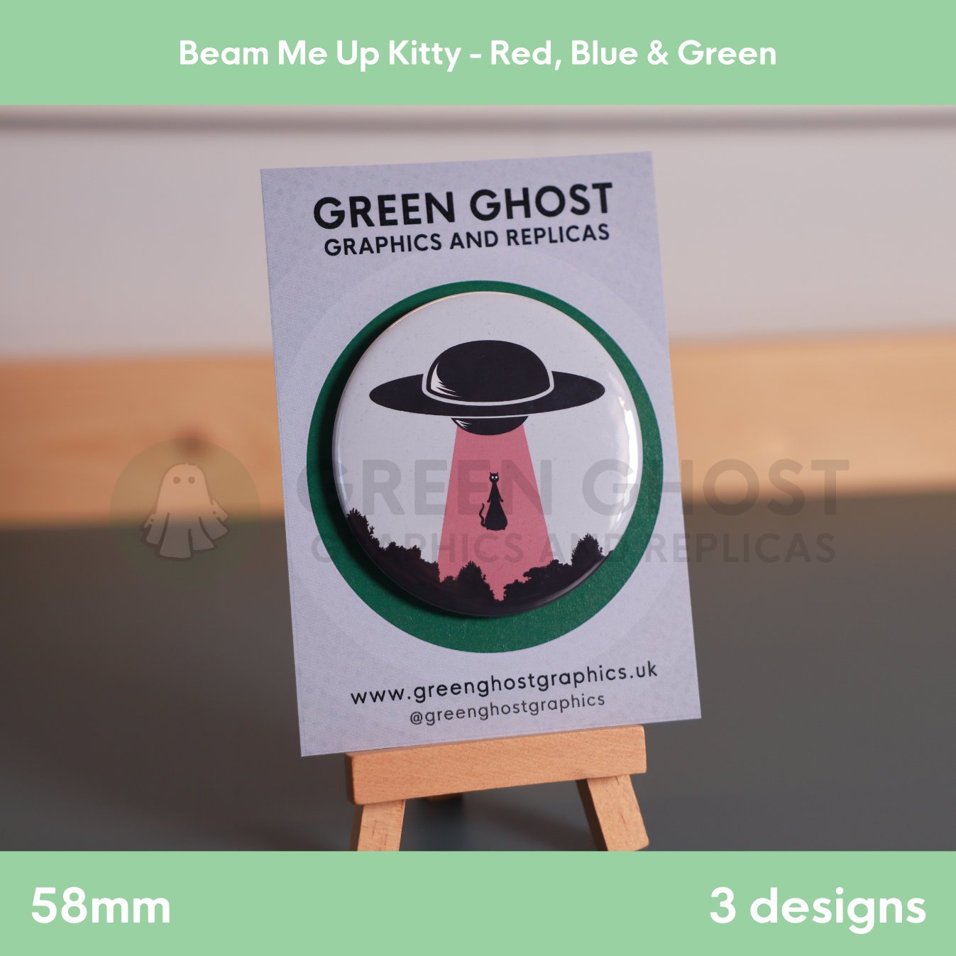 A round badge with a red UFO beam lifting a cat displayed on a branded backing card on a wooden easel.