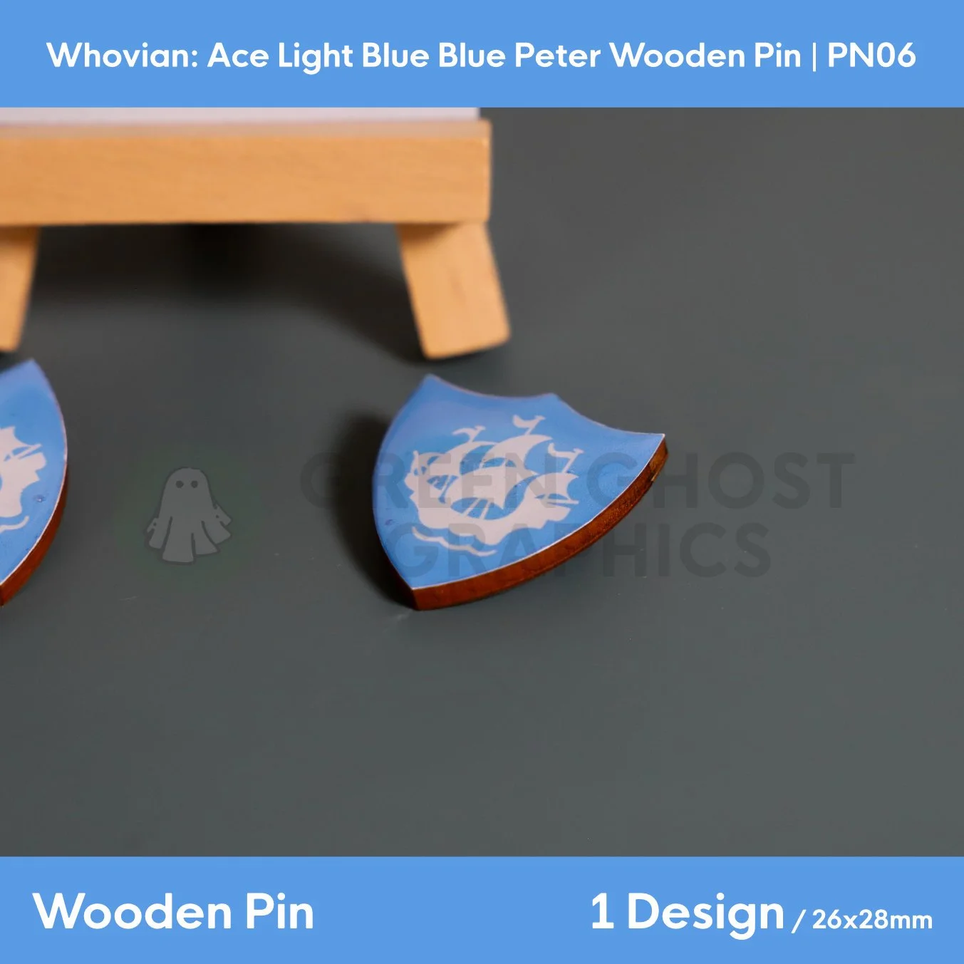 A blue shield‑shaped wooden pin with a white ship emblem shown lying flat on a smooth grey surface.