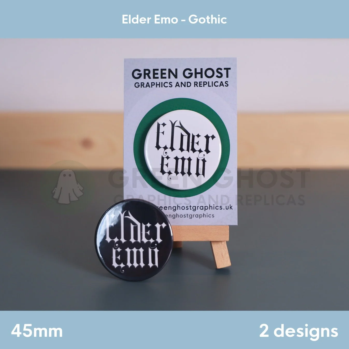 Two round badges, one black and one white, with "Elder Emo" text displayed together on a wooden easel with a branded backing card behind them.