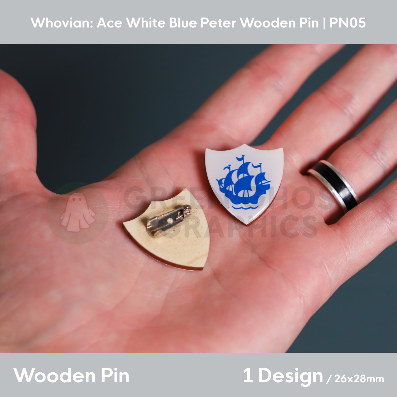 A white shield‑shaped wooden pin featuring a blue sailing ship emblem shown in a hand alongside the natural wood reverse with a metal brooch fastening.