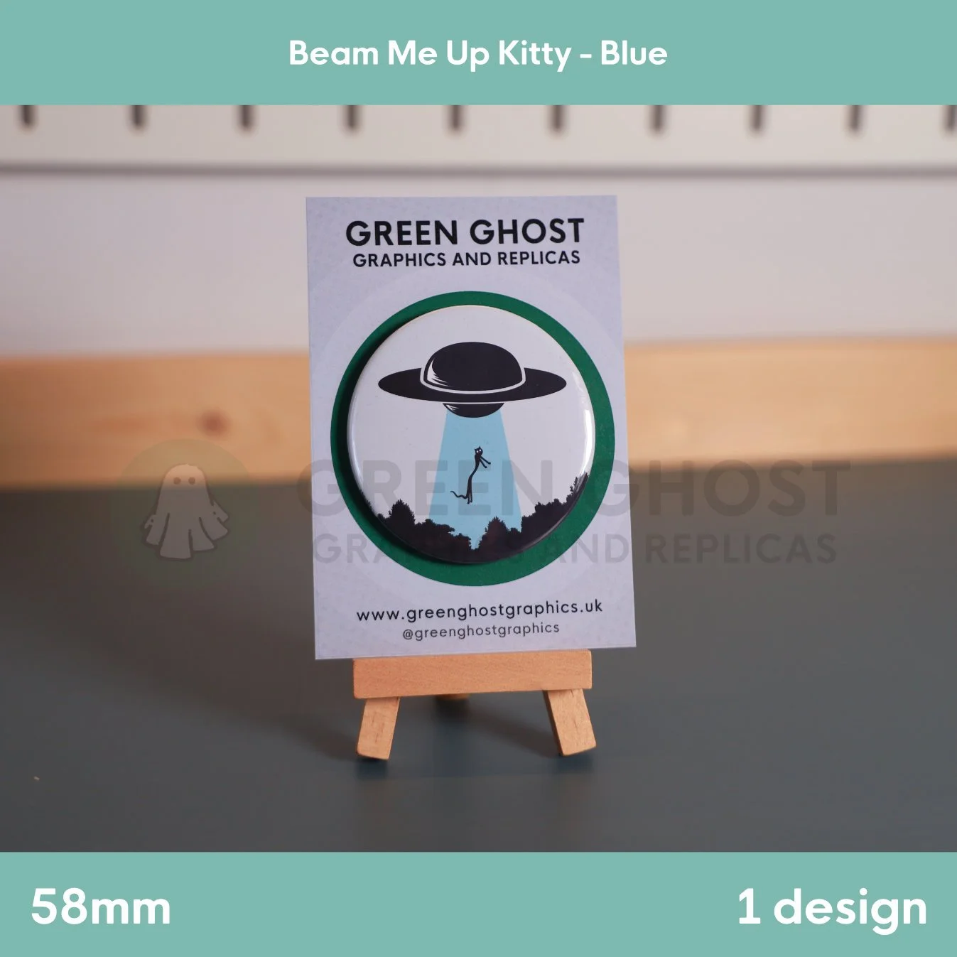 A round badge with a blue UFO beam lifting a cat displayed on a branded backing card on a wooden easel.