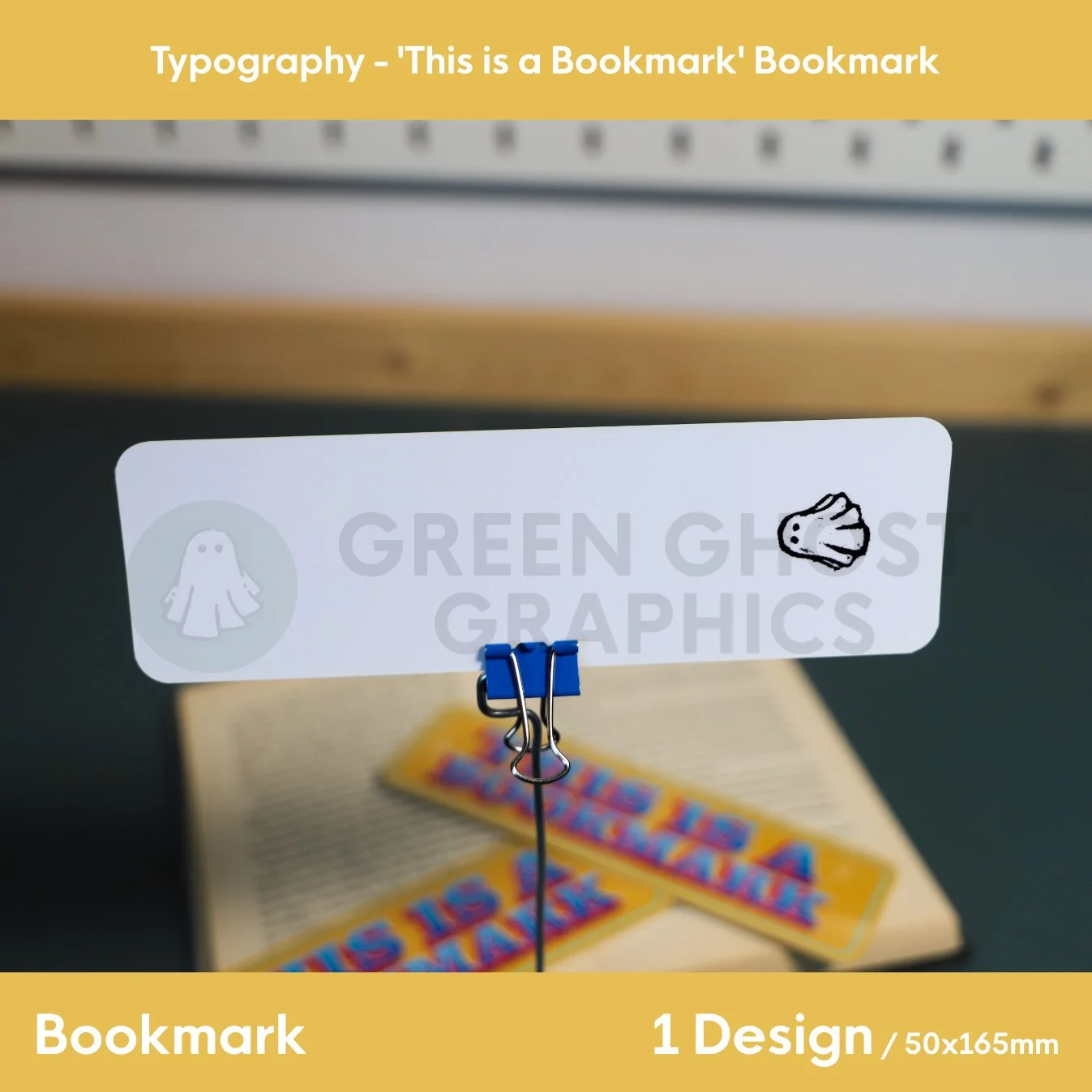 A bright yellow bookmark printed with large blue and red “THIS IS A BOOKMARK” text displayed upright on a metal stand.