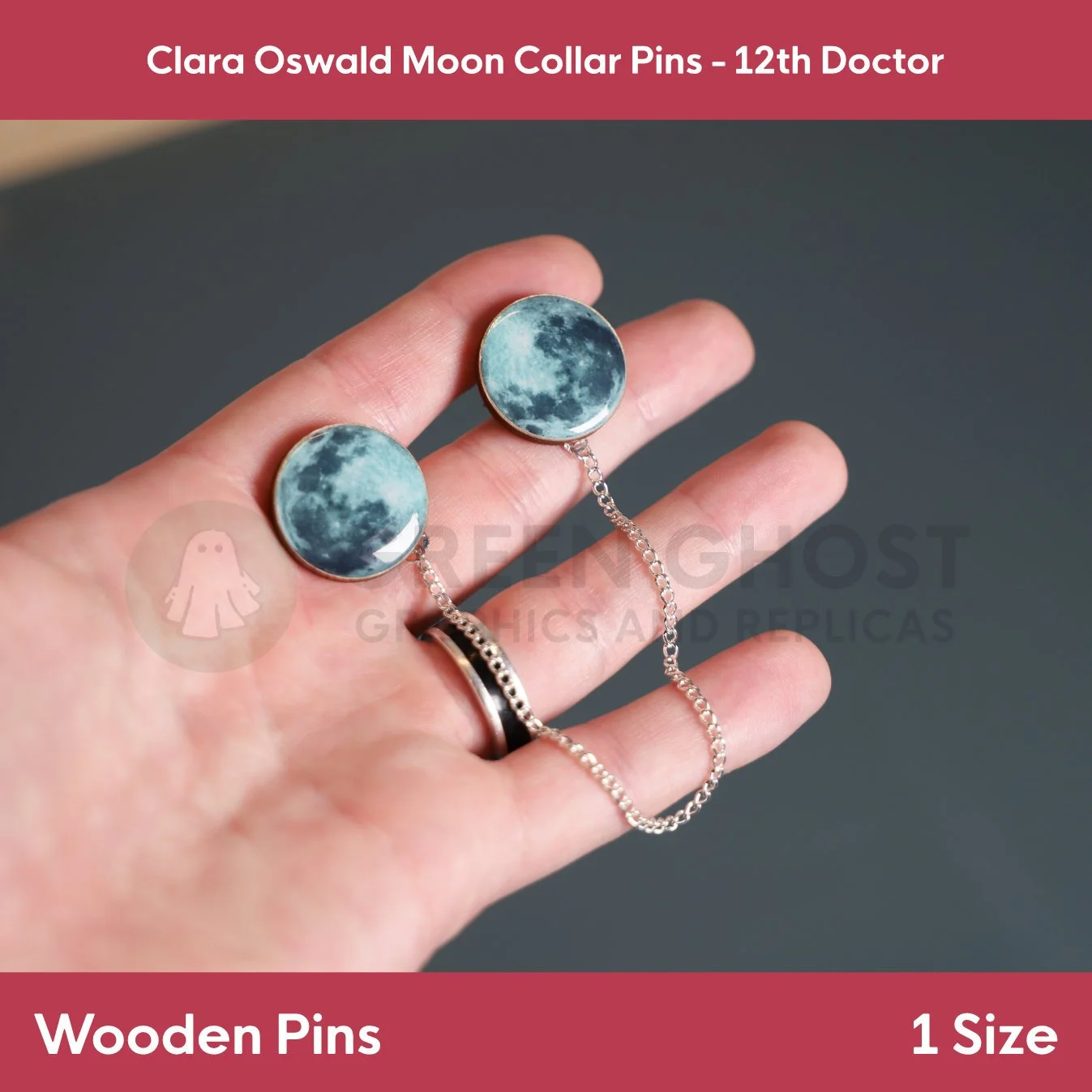 Two round moon‑image pins linked by a silver chain held in a hand against a neutral background.