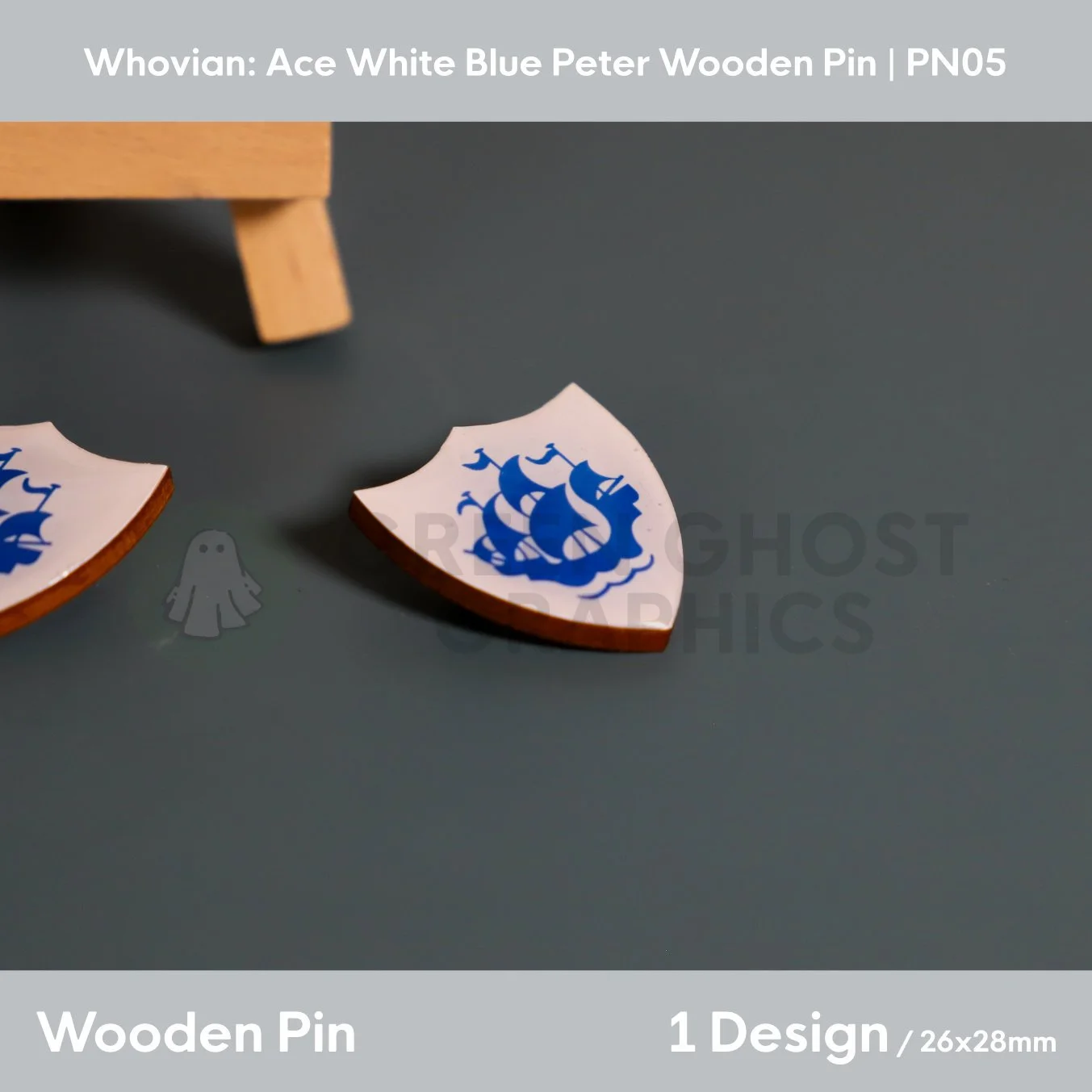 A white shield‑shaped wooden pin with a blue ship emblem shown lying flat on a smooth grey surface.