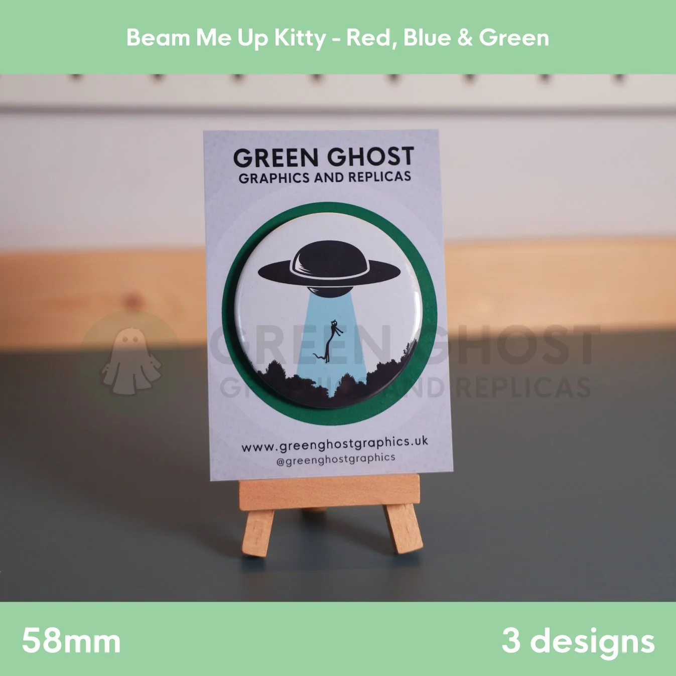 A round badge with a blue UFO beam lifting a cat displayed on a branded backing card on a wooden easel.