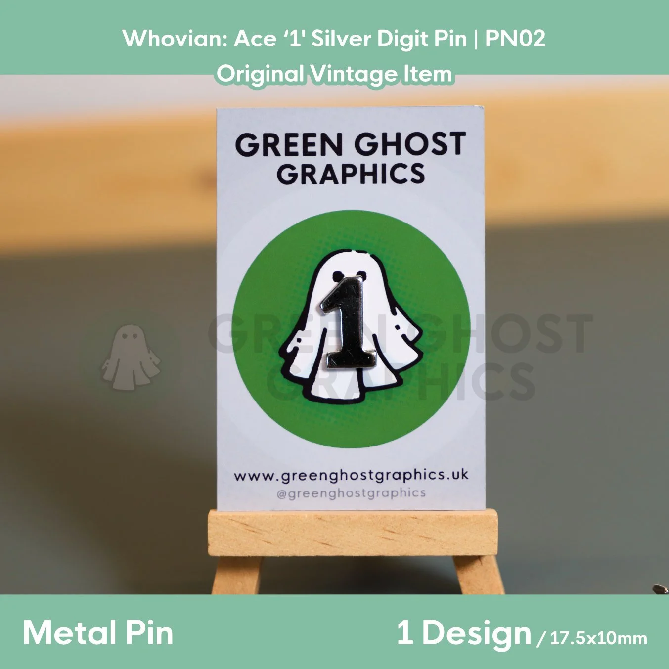A silver metal pin shaped like the number “1” mounted on a Green Ghost Graphics display card resting on a small wooden easel.