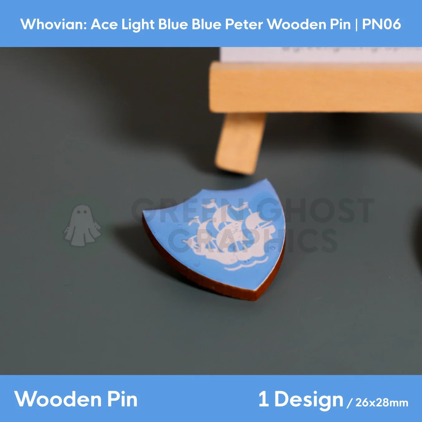 A blue shield‑shaped wooden pin featuring a white sailing ship design displayed on a dark surface beside a wooden easel.