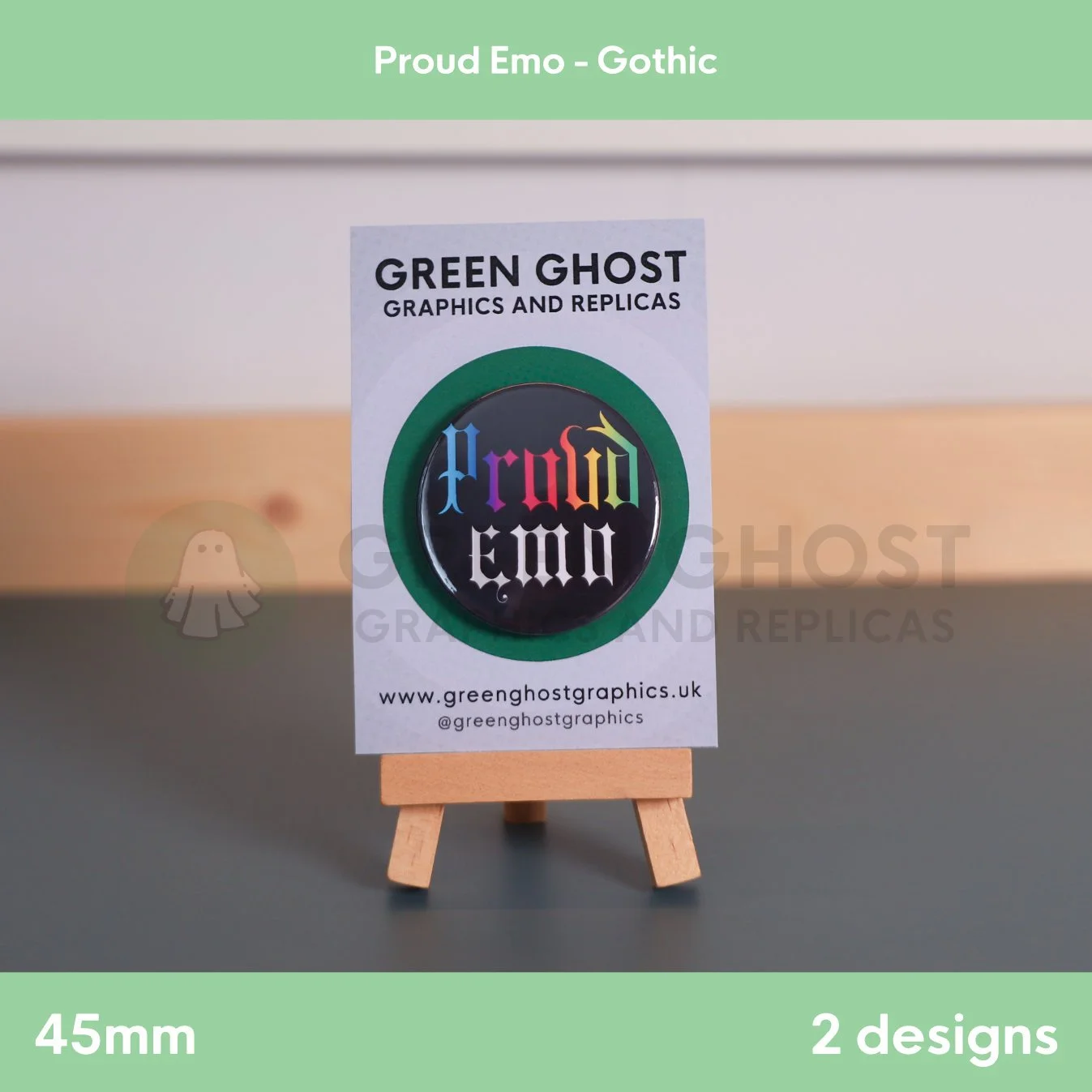 A round black badge with multicoloured “Proud Emo” text mounted on a branded backing card on a wooden easel.