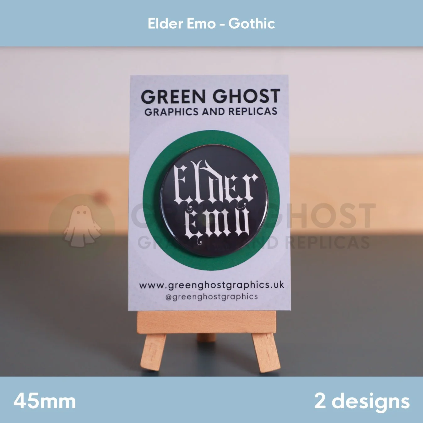 A round black badge with white Gothic "Elder Emo" text displayed on a branded backing card on a wooden easel.