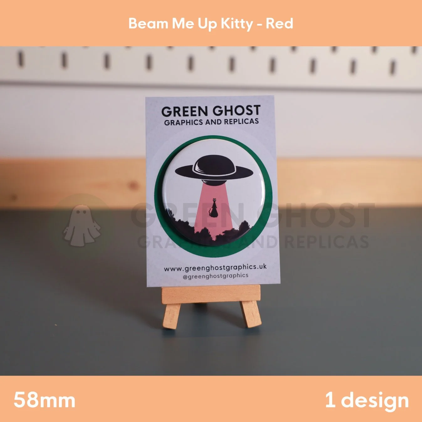 A round badge with a black ufo and red UFO beam lifting a cat displayed on a branded backing card on a wooden easel.