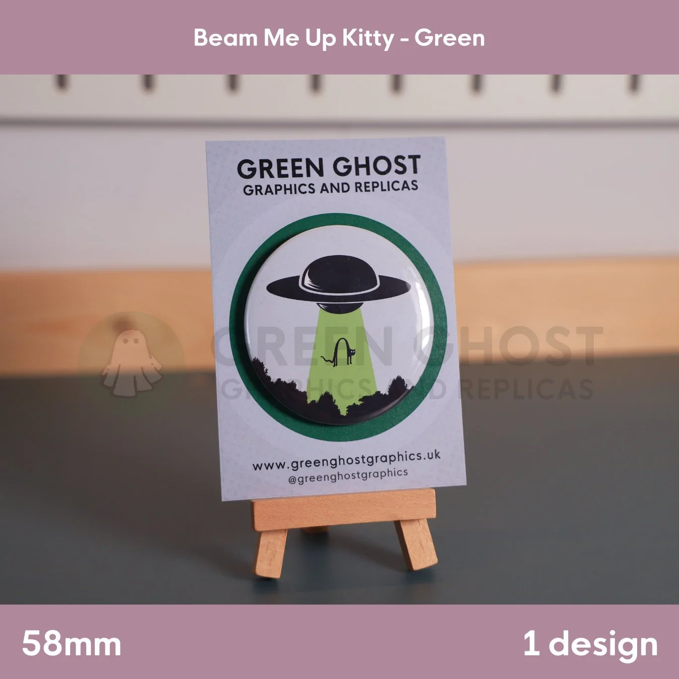 A round badge with a green UFO beam lifting a cat displayed on a branded backing card on a wooden easel.