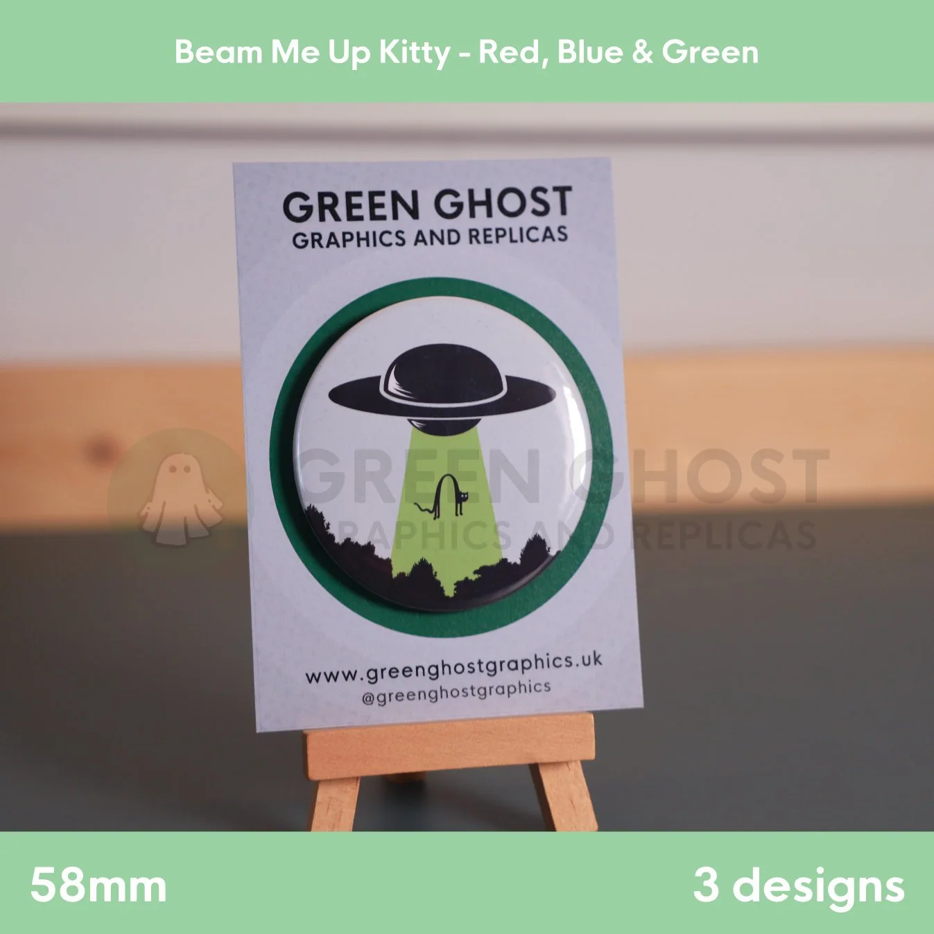 A round badge with a green UFO beam lifting a cat displayed on a branded backing card on a wooden easel.
