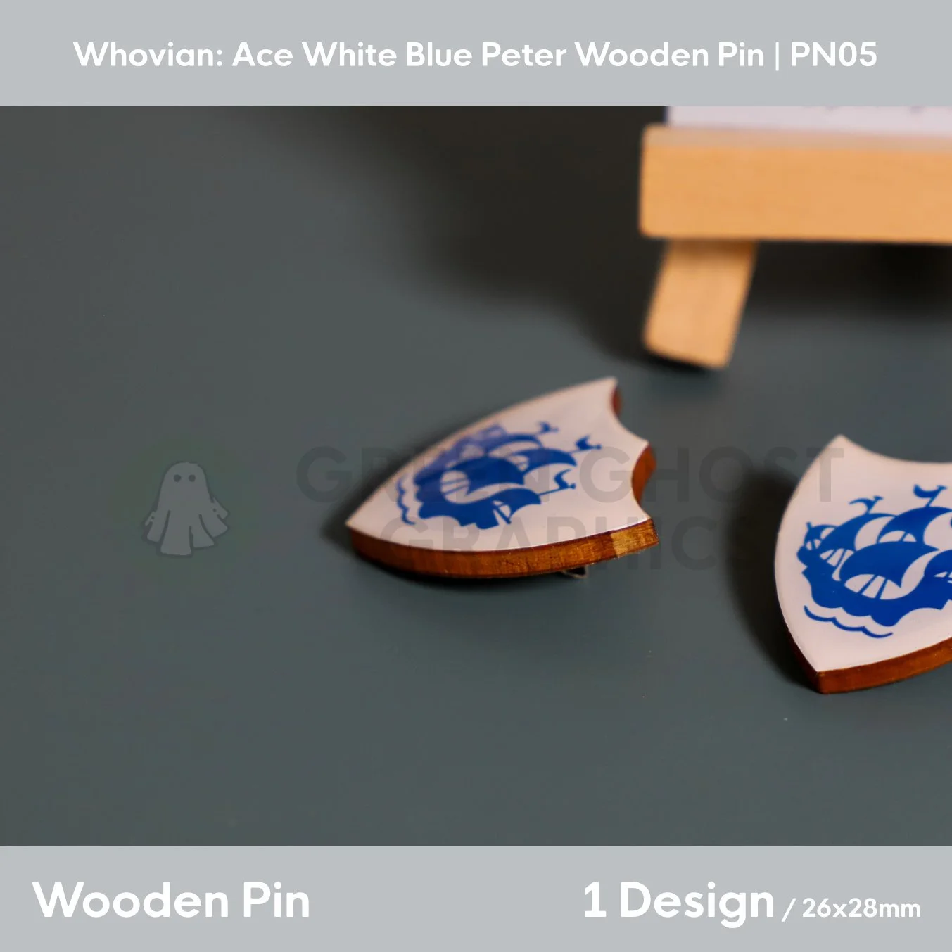 A white shield‑shaped wooden pin featuring a blue sailing ship design displayed on a dark surface beside a wooden easel.