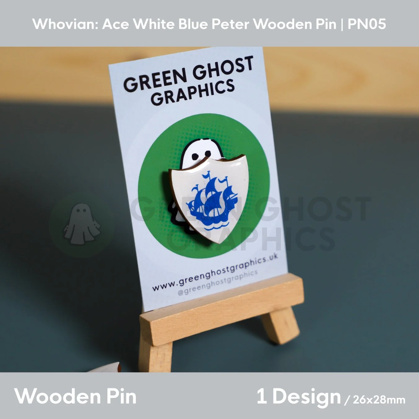 A white shield‑shaped wooden pin displaying a blue sailing ship motif mounted on a Green Ghost Graphics card on a small wooden easel.