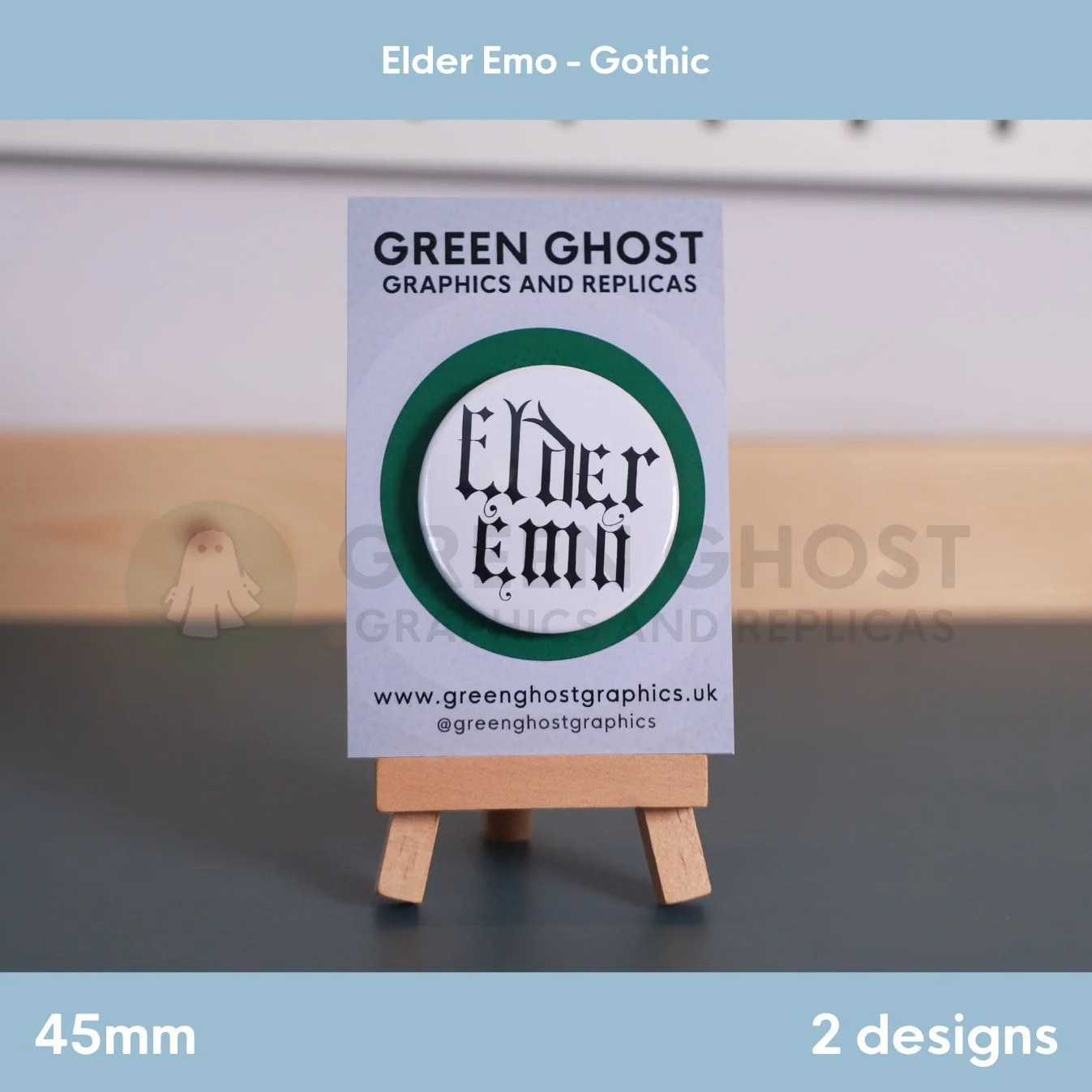 A round white badge with black Gothic "Elder Emo" text displayed on a branded backing card on a wooden easel.