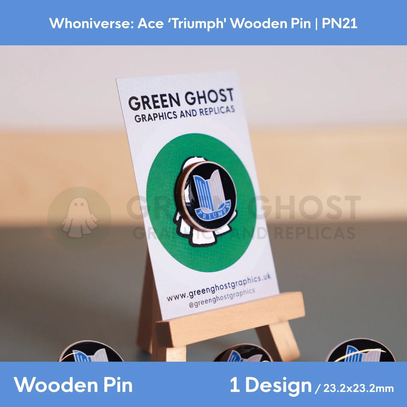A circular wooden pin featuring a blue and white Triumph emblem, attached to a branded backing card on a wooden easel, with extra pins visible in the background.