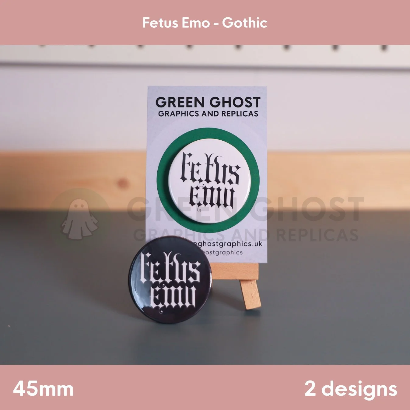 Two round badges, one black and one white, with "Fetus Emo" text displayed together on a wooden easel with a branded backing card behind them.