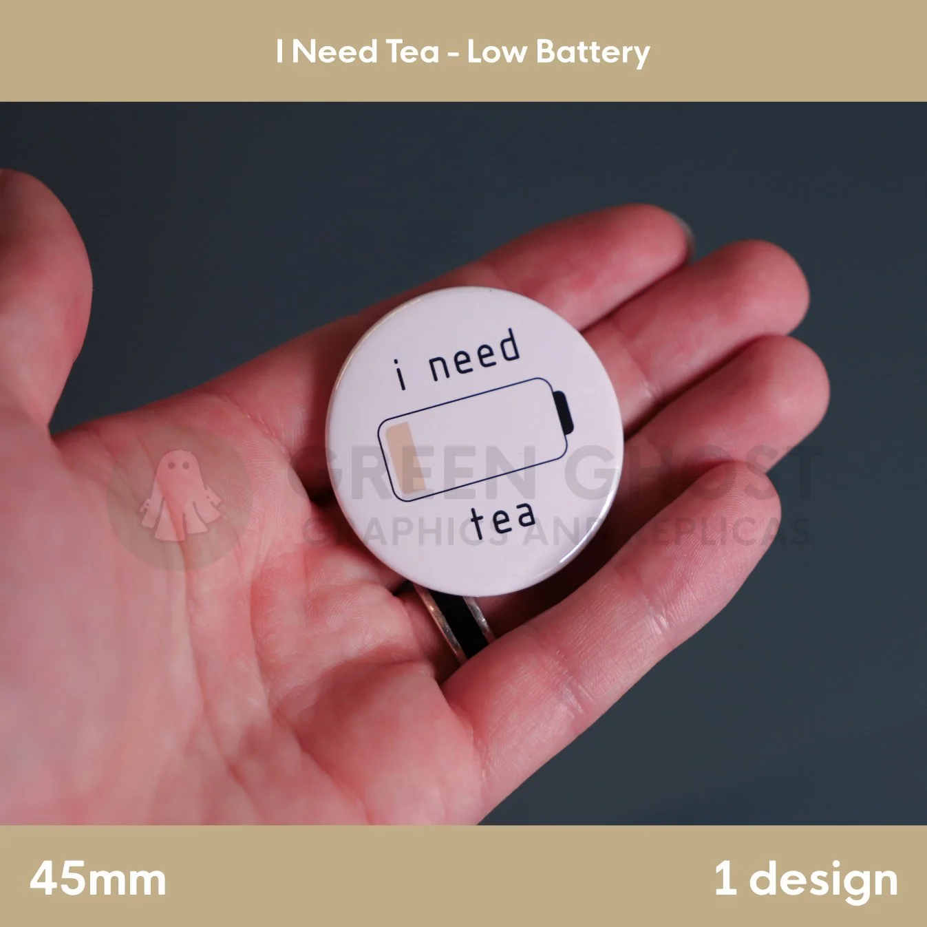 A round white badge with a low‑battery icon and the phrase “i need tea” held in a hand against a grey background.