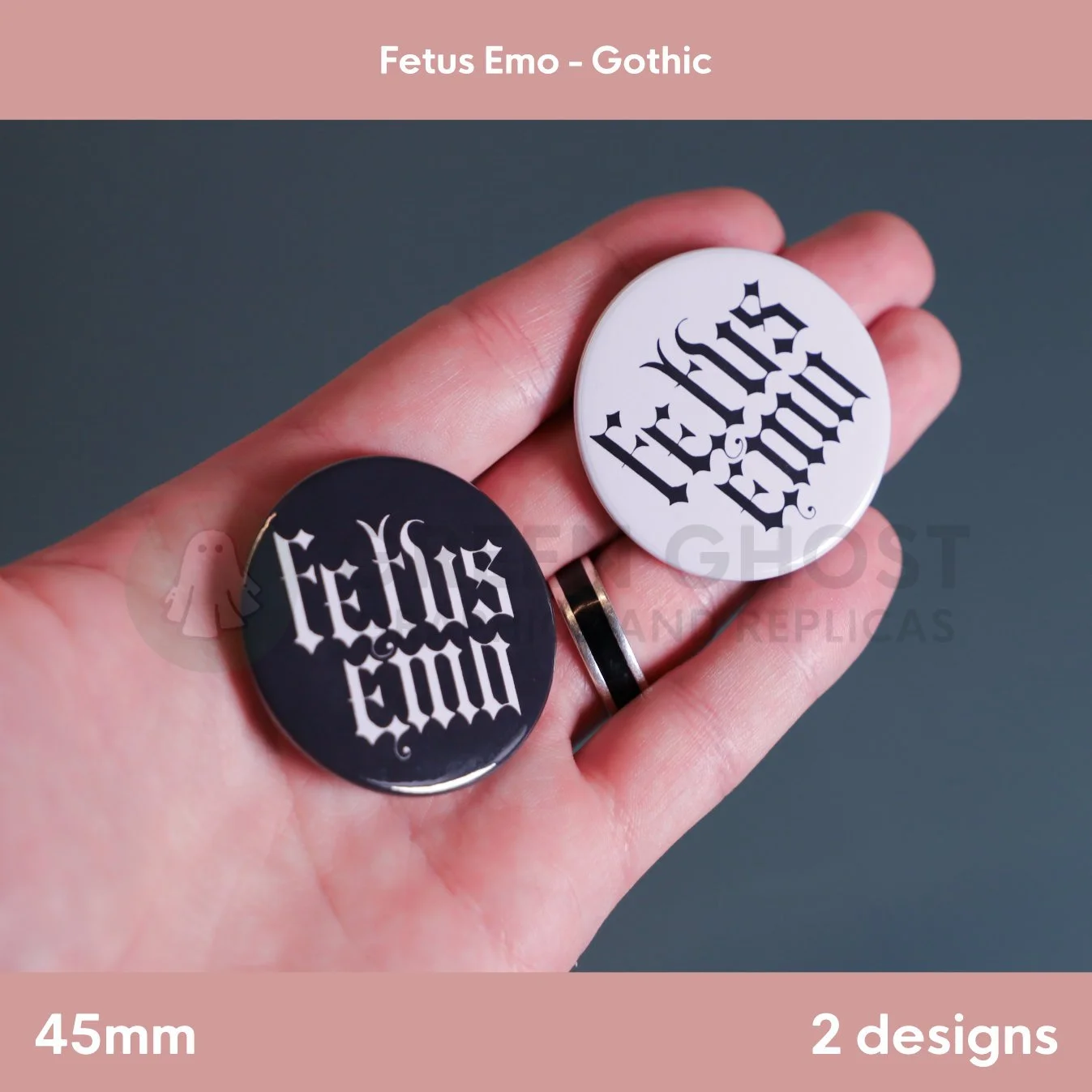 A hand holds two round badges, one black with white Gothic "Fetus Emo" text and one white with black Gothic text, against a muted background.
