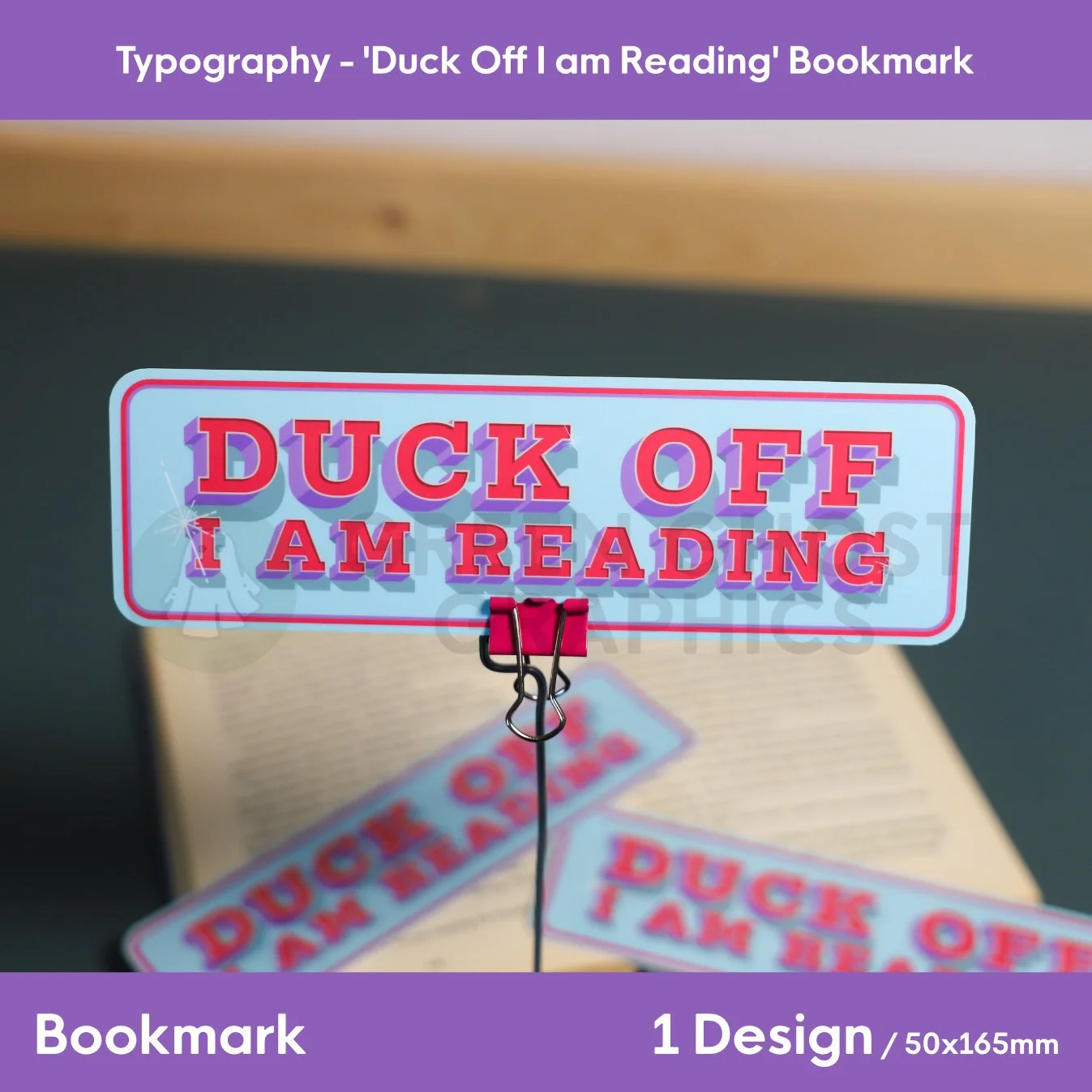A rectangular light‑blue bookmark with bold red and purple “DUCK OFF I AM READING” text held upright in a binder clip beside an open book.