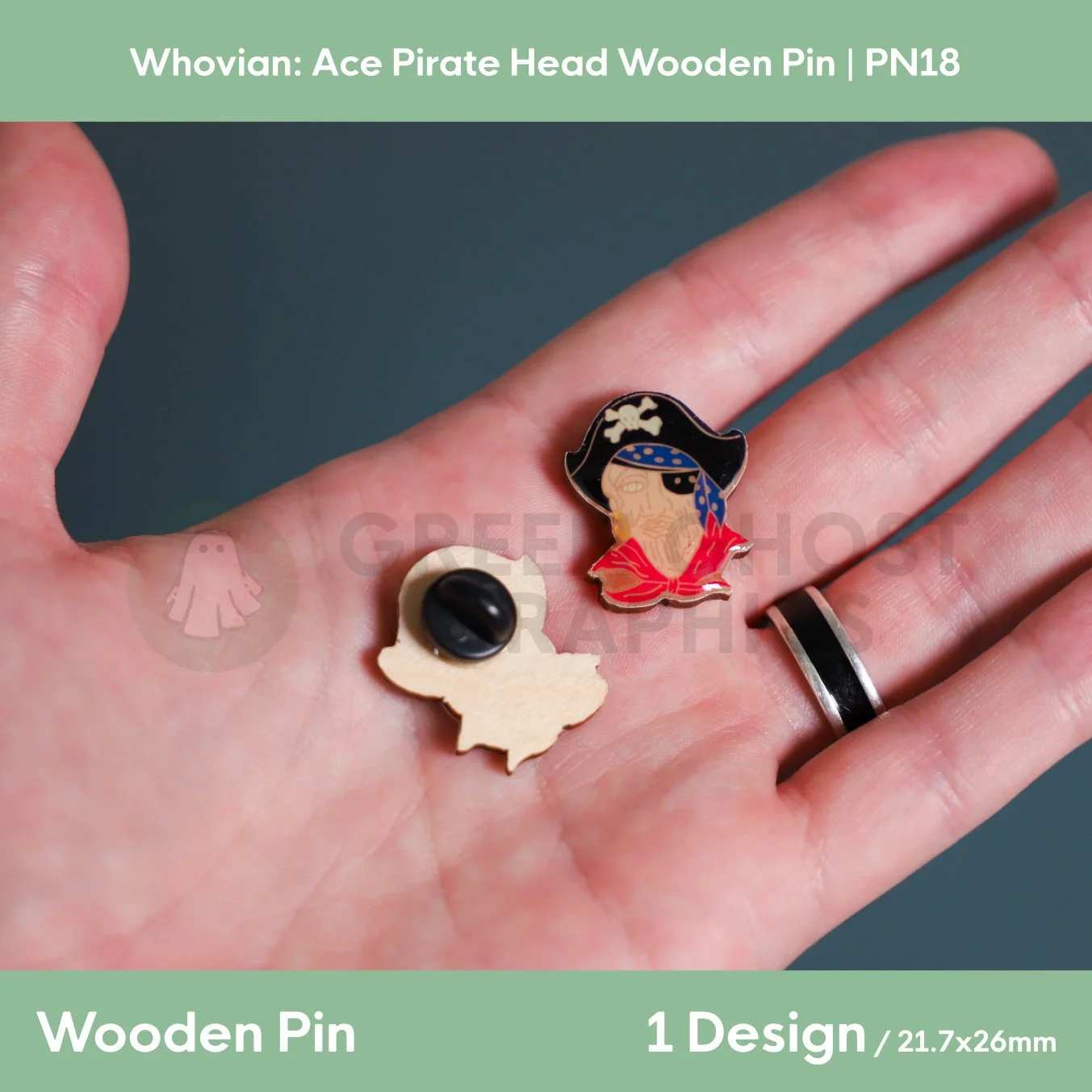 A pirate‑head wooden pin with a black pirate hat and red clothing displayed in a hand beside the natural wood reverse with a black rubber clasp.