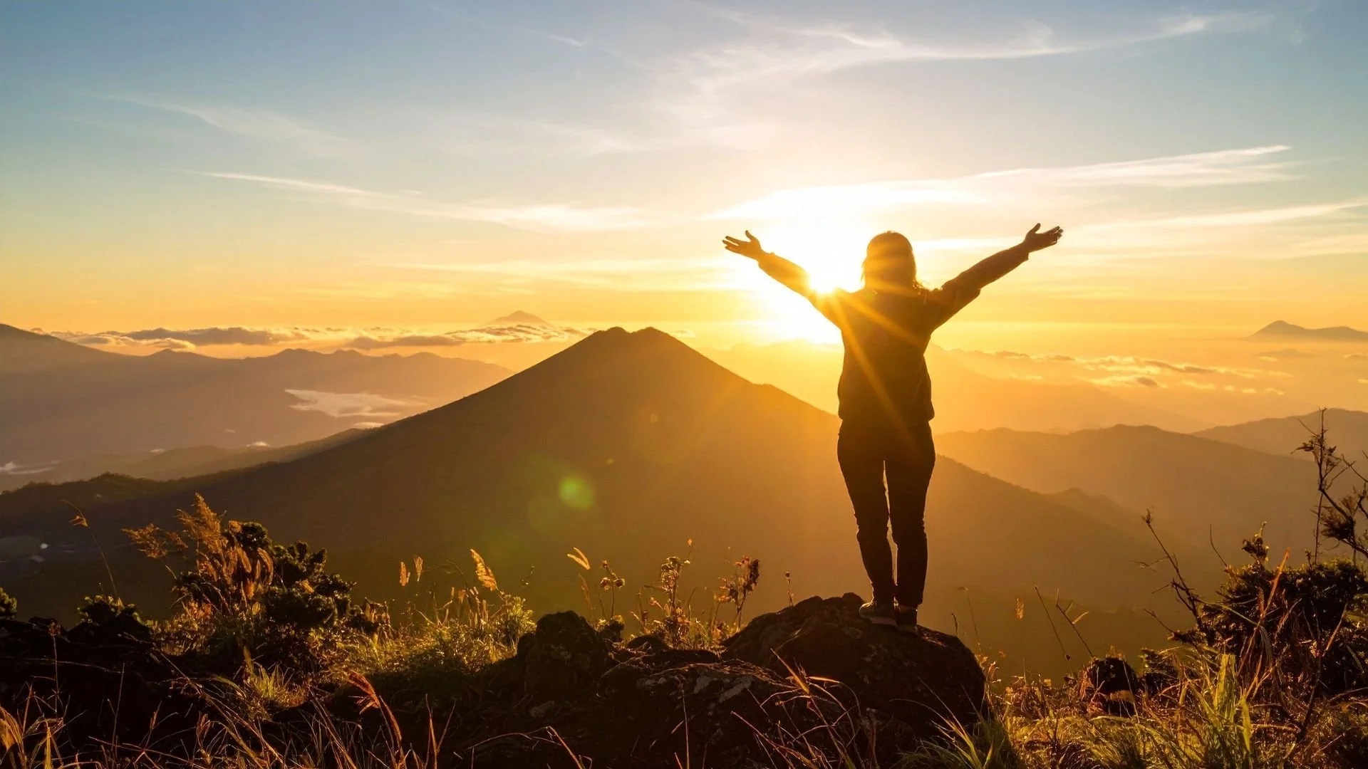 Person standing on a mountain with arms outstretched during sunrise, overlooking distant mountains and clouds.
